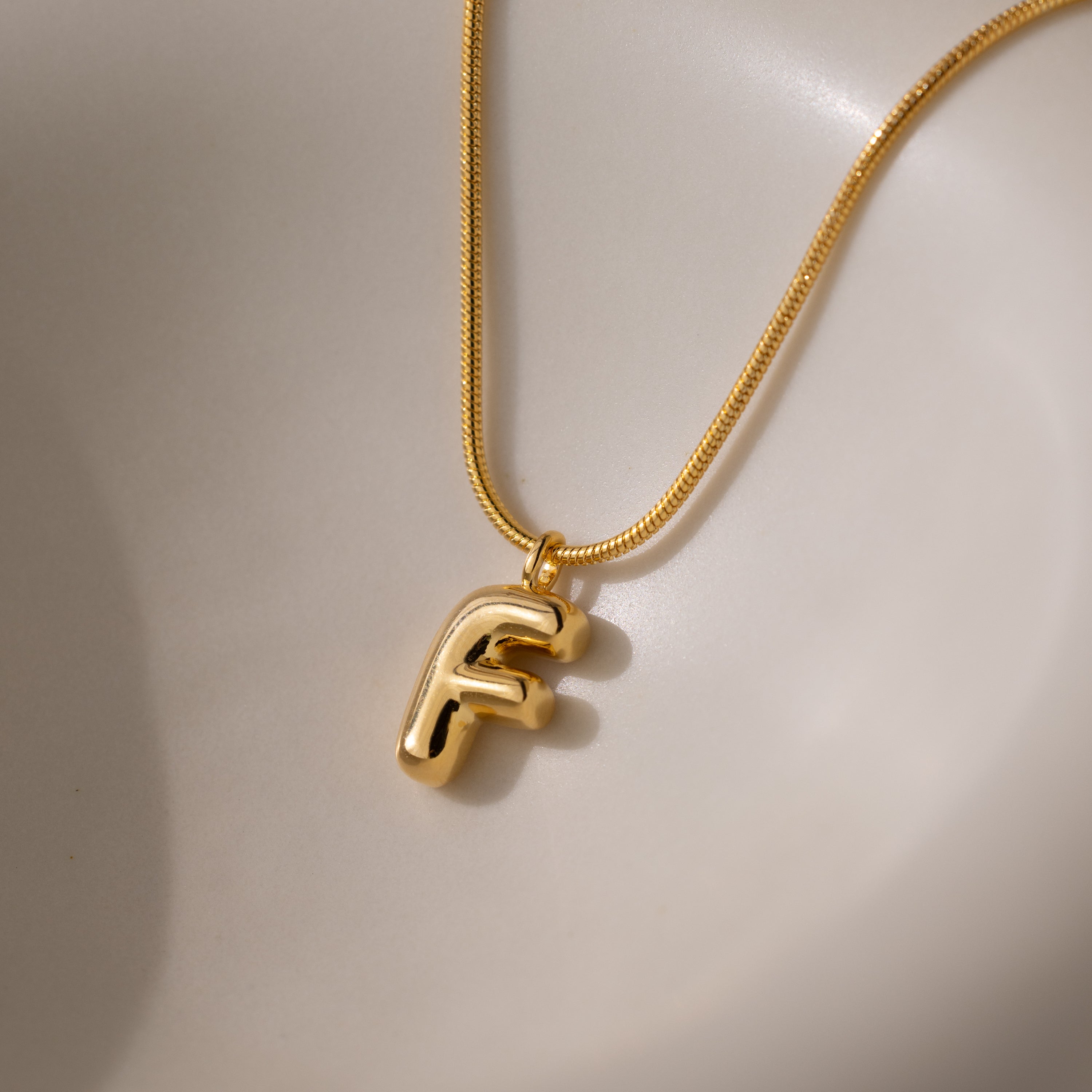 The Bubble Letter Necklace in 18K Gold features a chunky letter F charm pendant resting on a smooth, light surface, offering a bold twist on personalized jewelry.