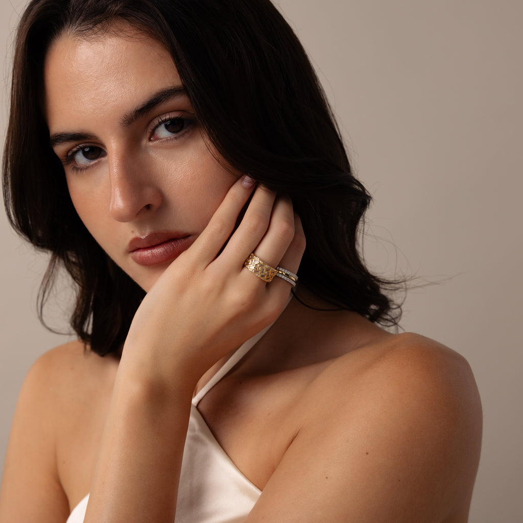 A woman with dark hair in a white top touches her face, showcasing the Mosaic Diamond Baguette Ring with gold accents as she looks at the camera.