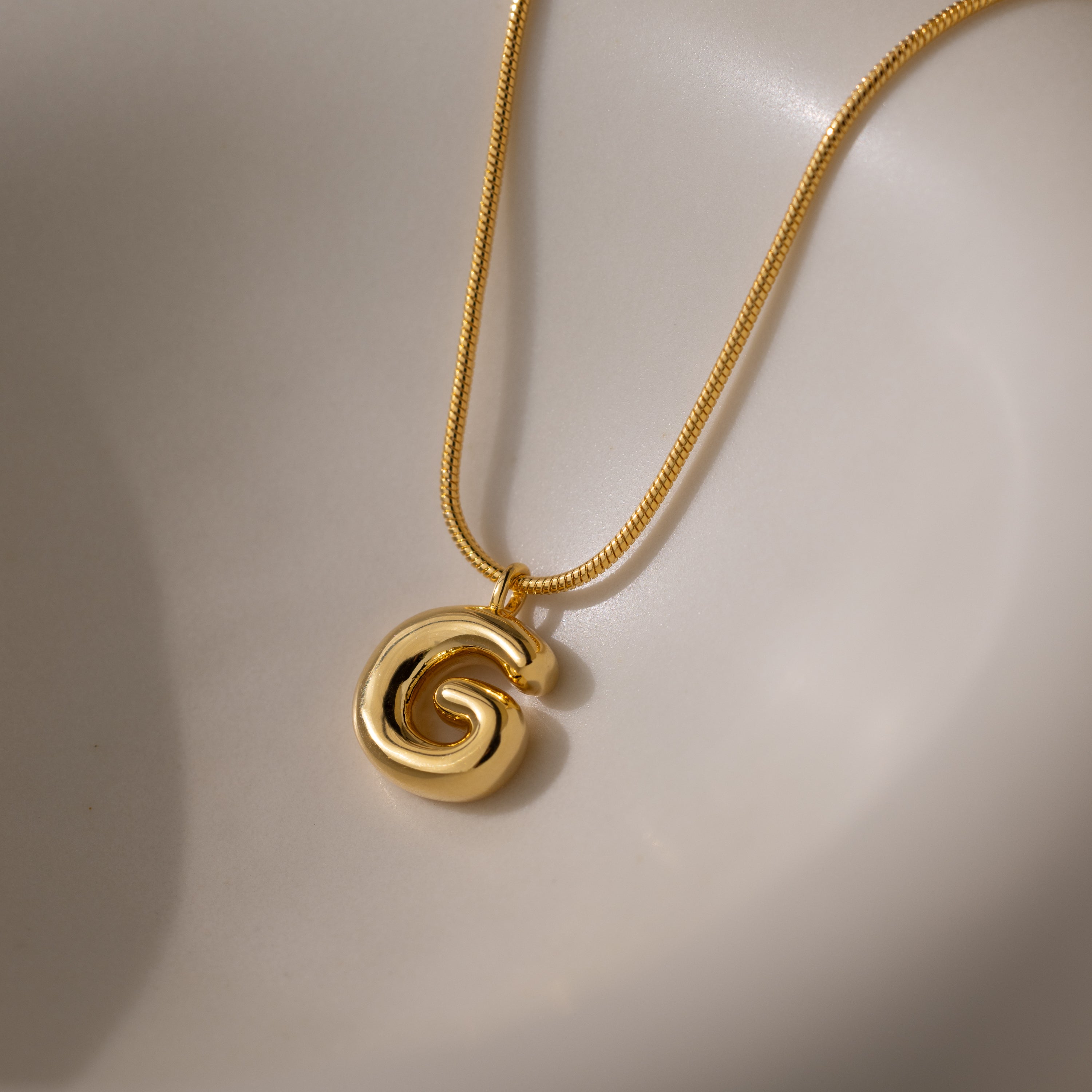 The Bubble Letter Necklace in 18K Gold features a polished spiral design, shown on a smooth, light surface—ideal for layering with chunky letter charms or mixing with personalized jewelry pieces.