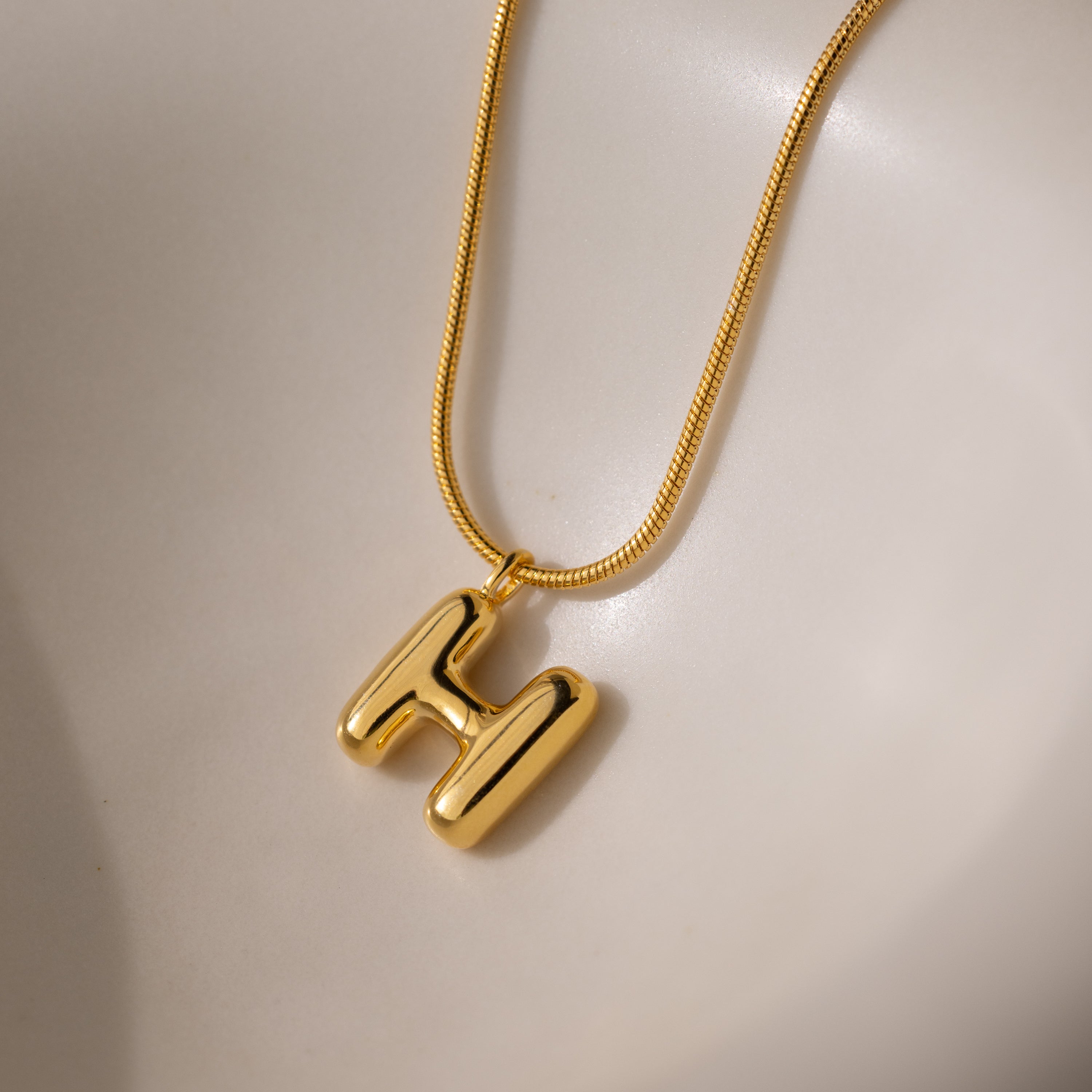 The Bubble Letter Necklace in 18K Gold features a shiny, chunky letter H pendant on a smooth, light-colored surface—a bold and personalized jewelry piece.
