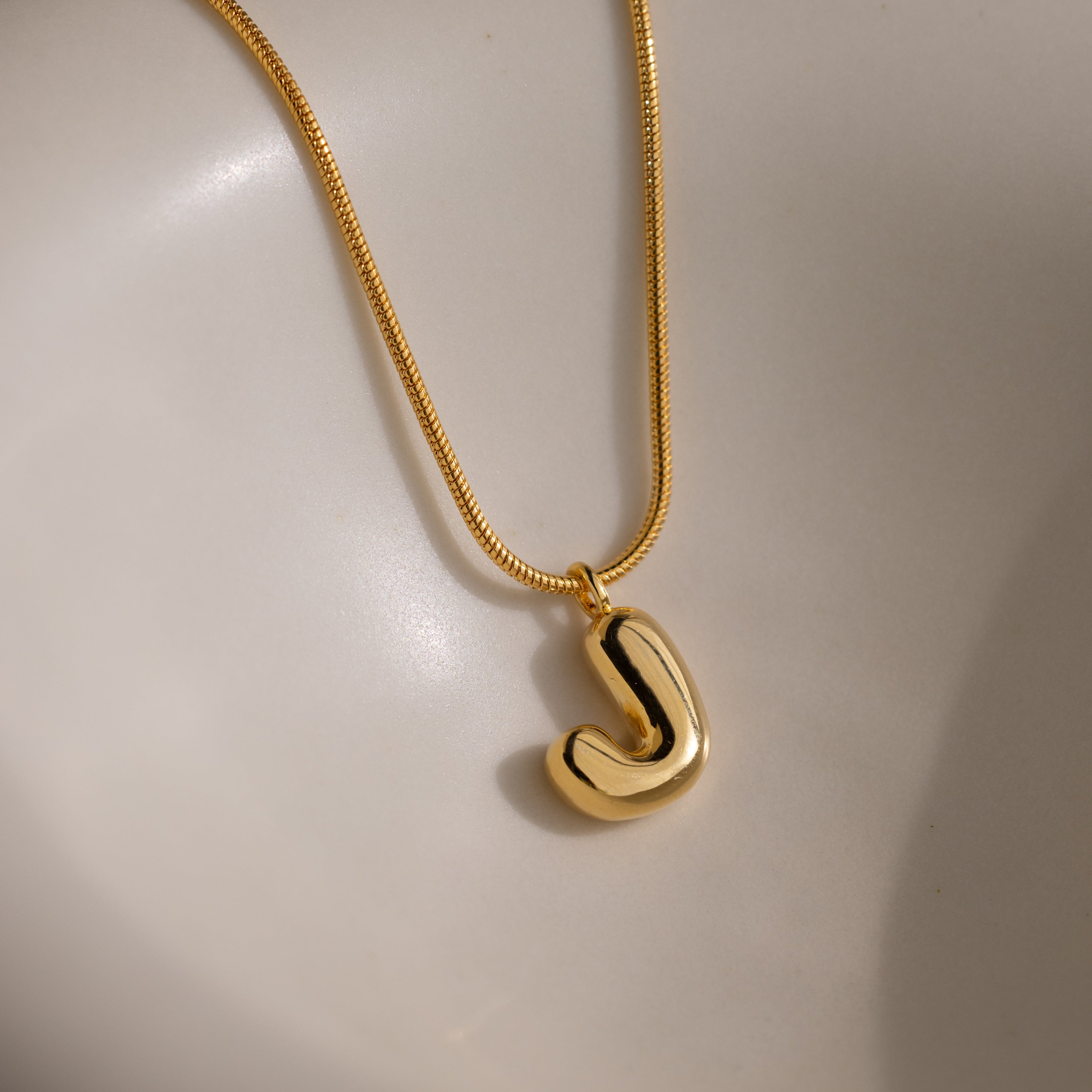 The Bubble Letter Necklace in 18K Gold features a shiny, chunky letter J pendant on a smooth background—ideal for anyone who loves personalized jewelry.