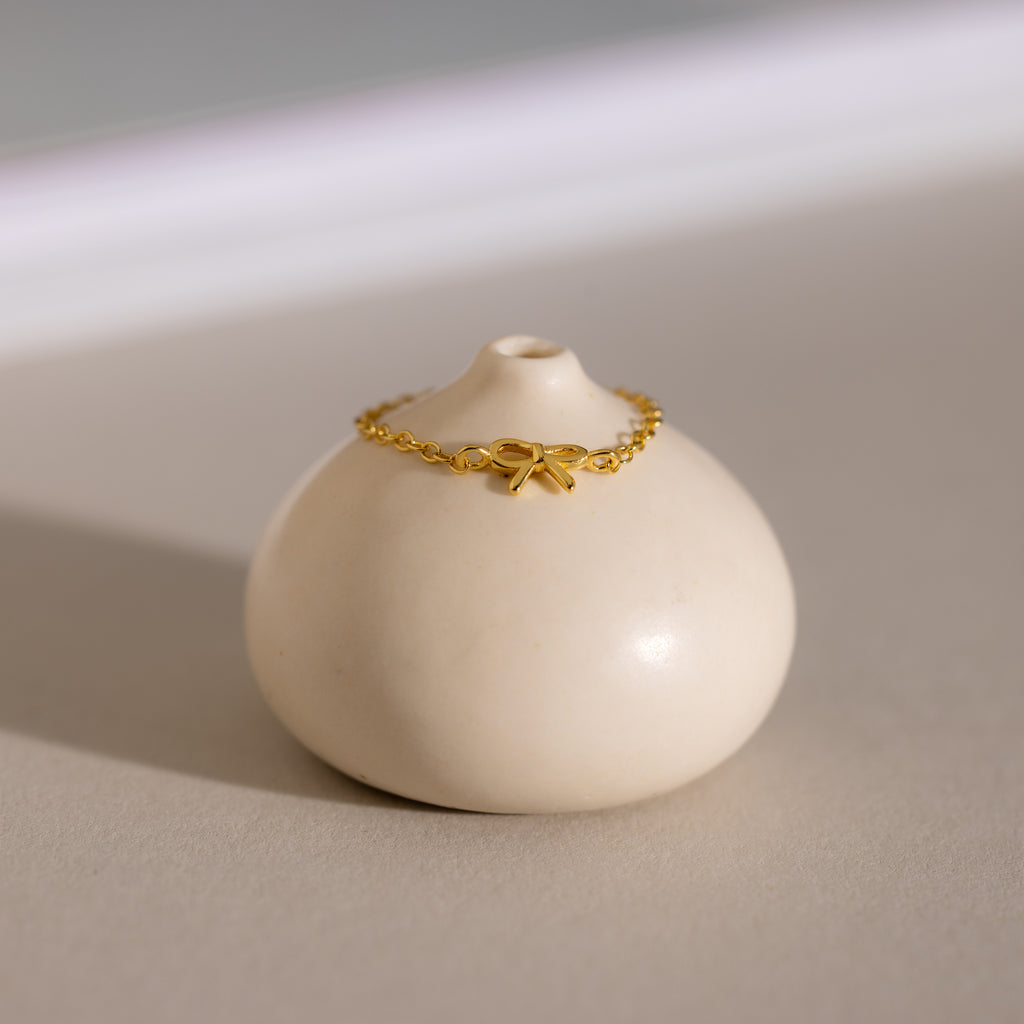 Delicate gold chain ring with a dainty bow charm displayed on a minimalist ceramic prop.