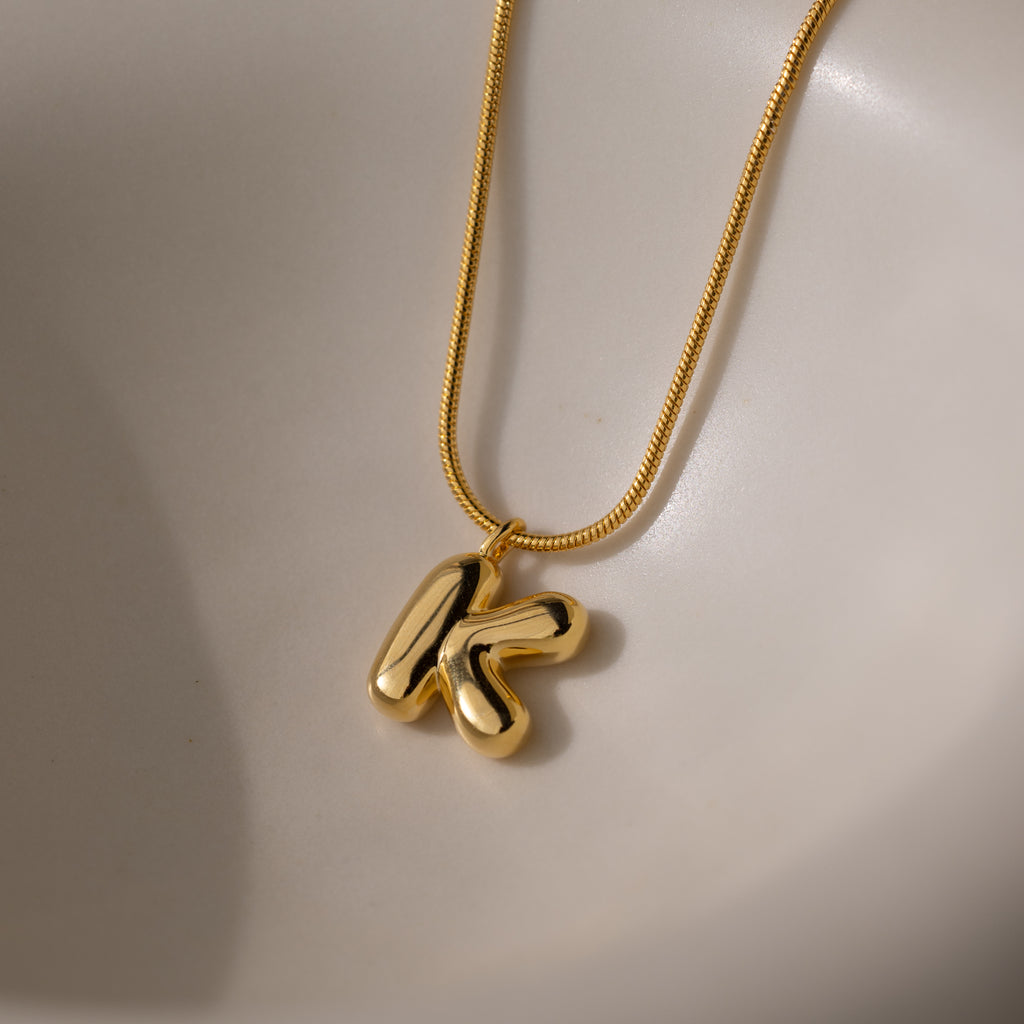 The Bubble Letter Necklace in 18K Gold features a shiny K pendant on a smooth, light surface, offering a playful and personalized jewelry accent.