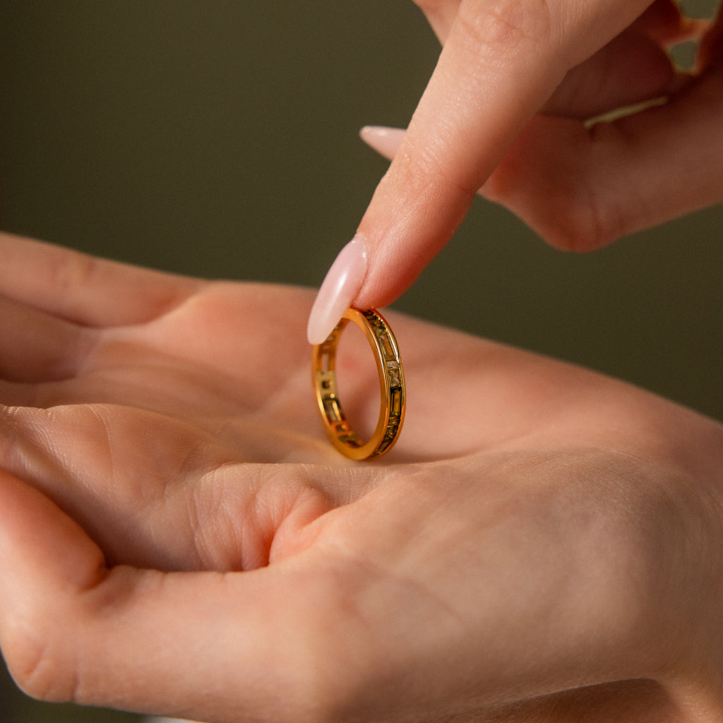 A hand holds the Peridot Inlay Eternity Ring as another hand gently touches it, set against a blurred background.