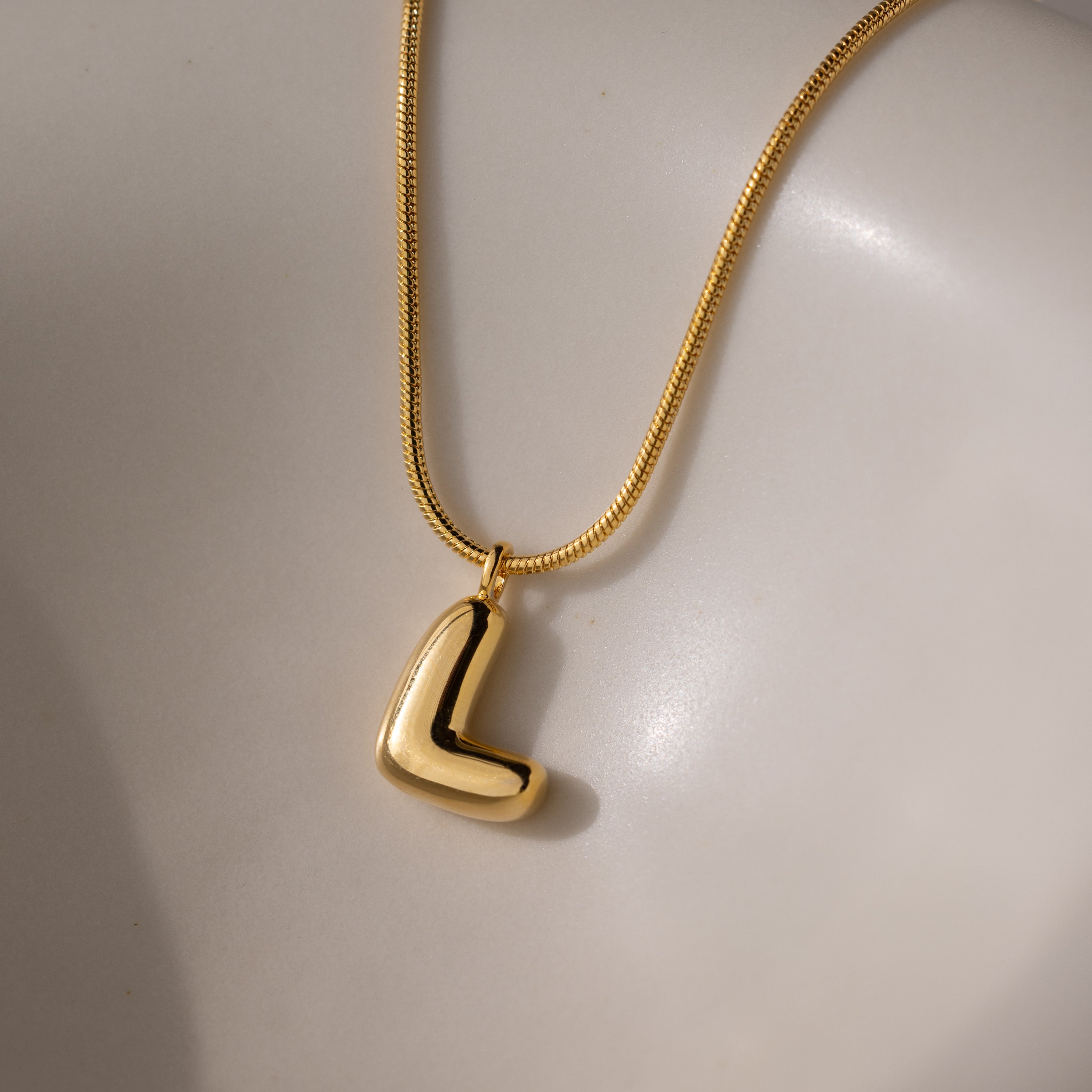 The Bubble Letter Necklace in 18K Gold, featuring a chunky L-shaped charm, rests on a smooth off-white surface.