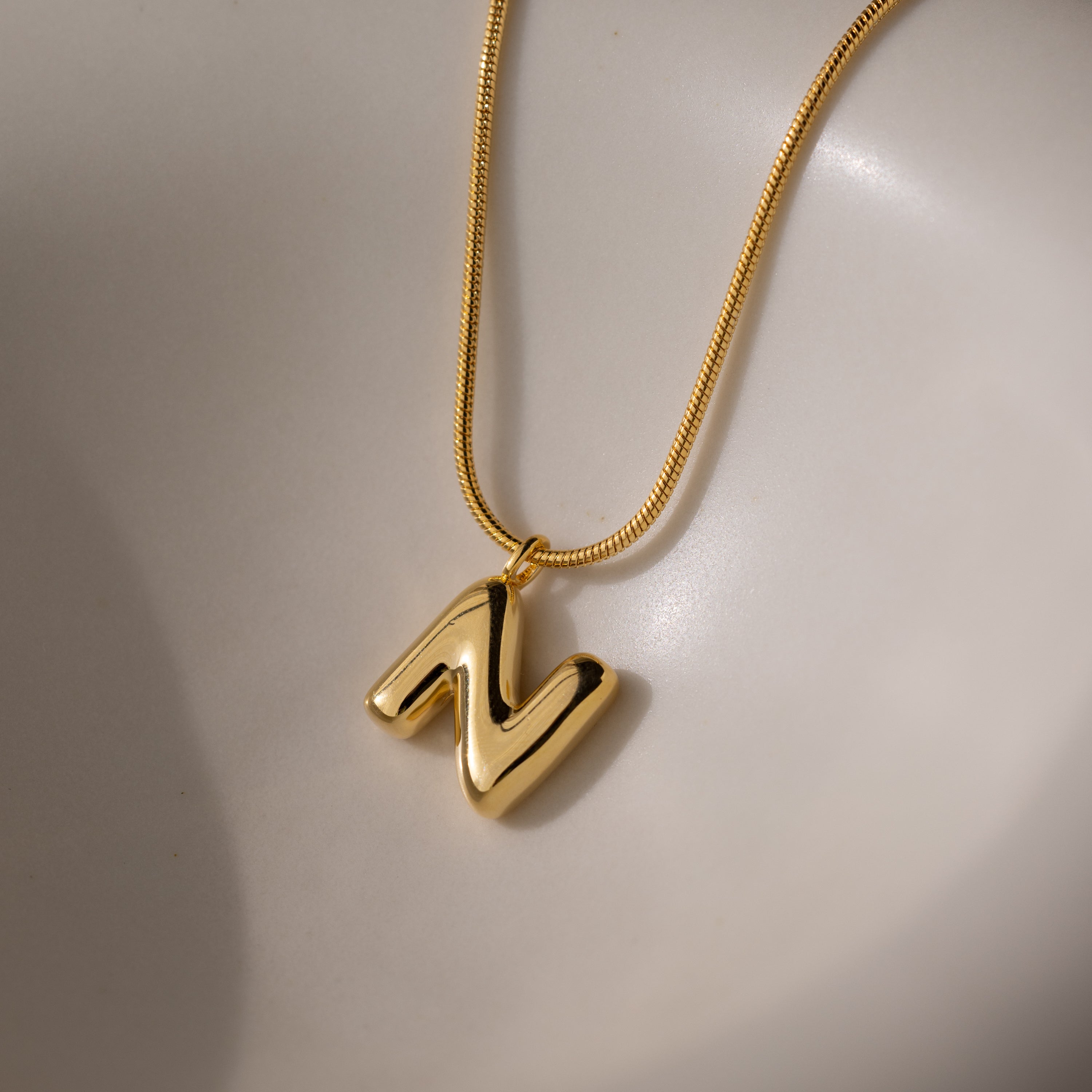 The Bubble Letter Necklace in 18K Gold features a bold N-shaped pendant on a smooth, light surface—a chic, personalized jewelry piece.