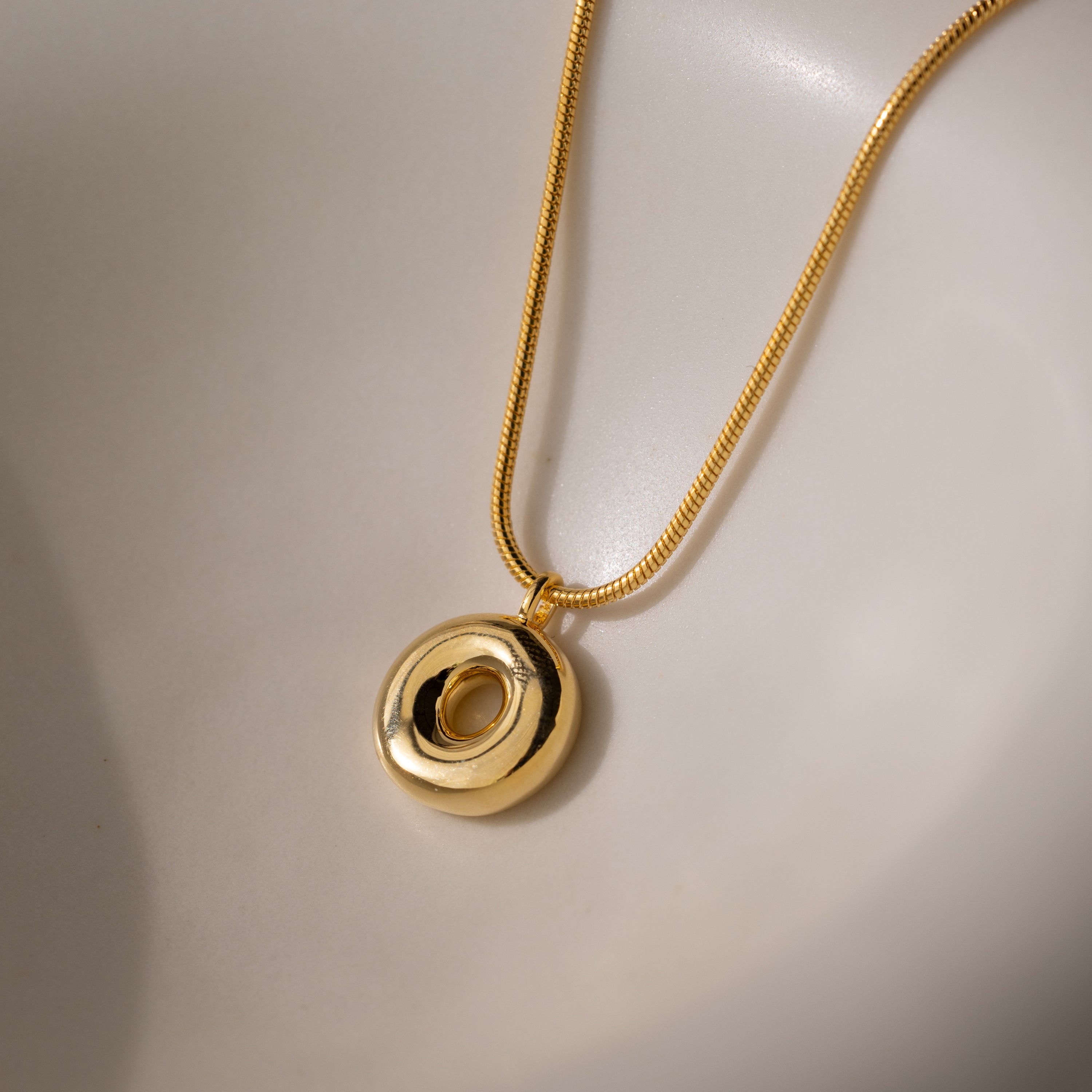 The Bubble Letter Necklace in 18K Gold features a smooth, circular design and rests on a white, curved surface—making it an elegant addition of personalized jewelry to any collection.