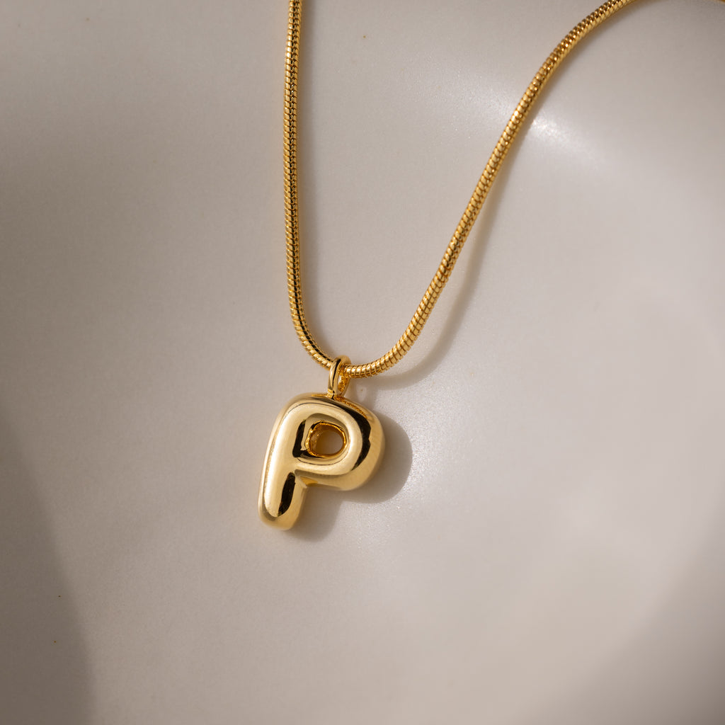 The Bubble Letter Necklace in 18K Gold showcases a shiny, chunky letter P charm pendant on a smooth, light surface—an elegant piece of personalized jewelry.