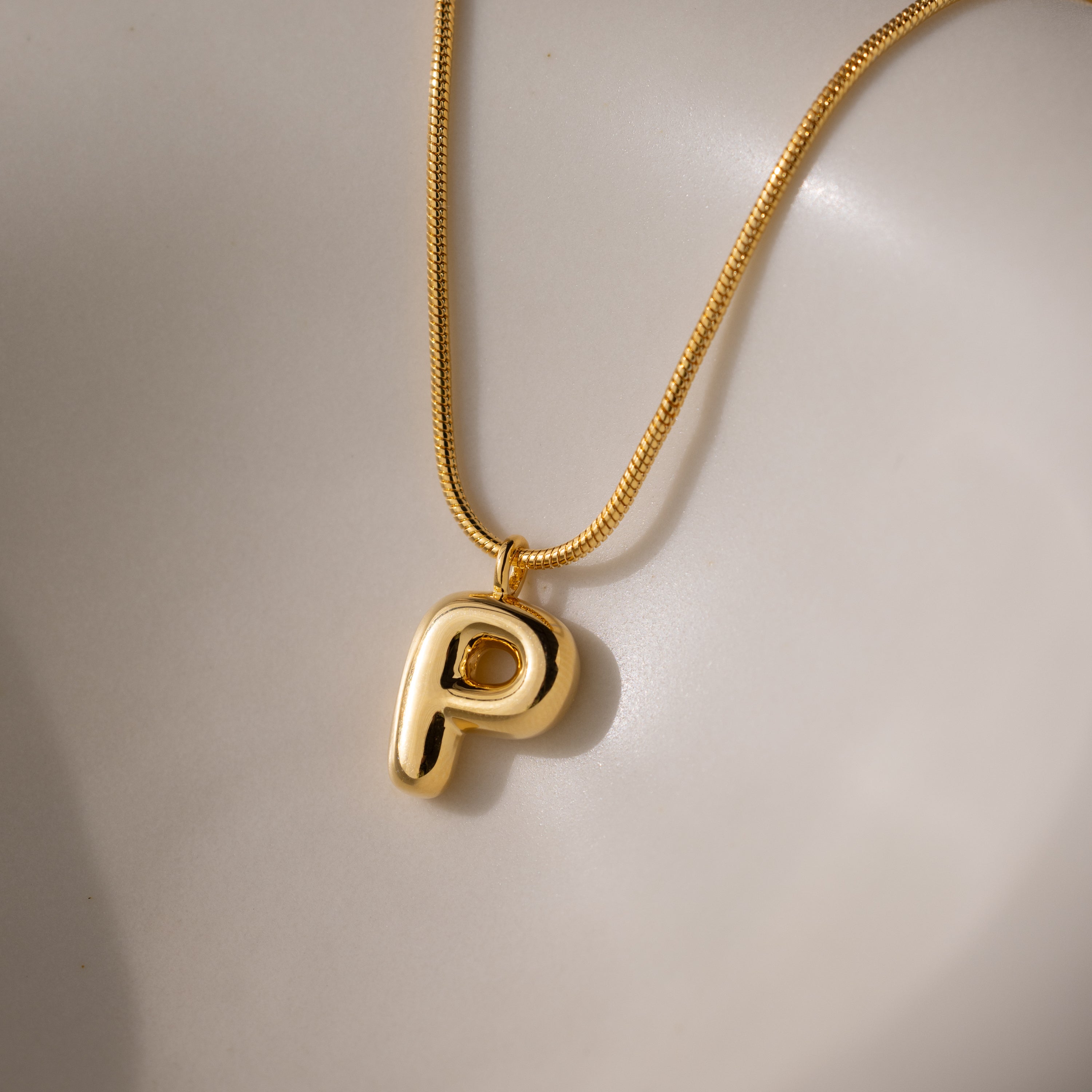 The Bubble Letter Necklace in 18K Gold showcases a shiny, chunky letter P charm pendant on a smooth, light surface—an elegant piece of personalized jewelry.