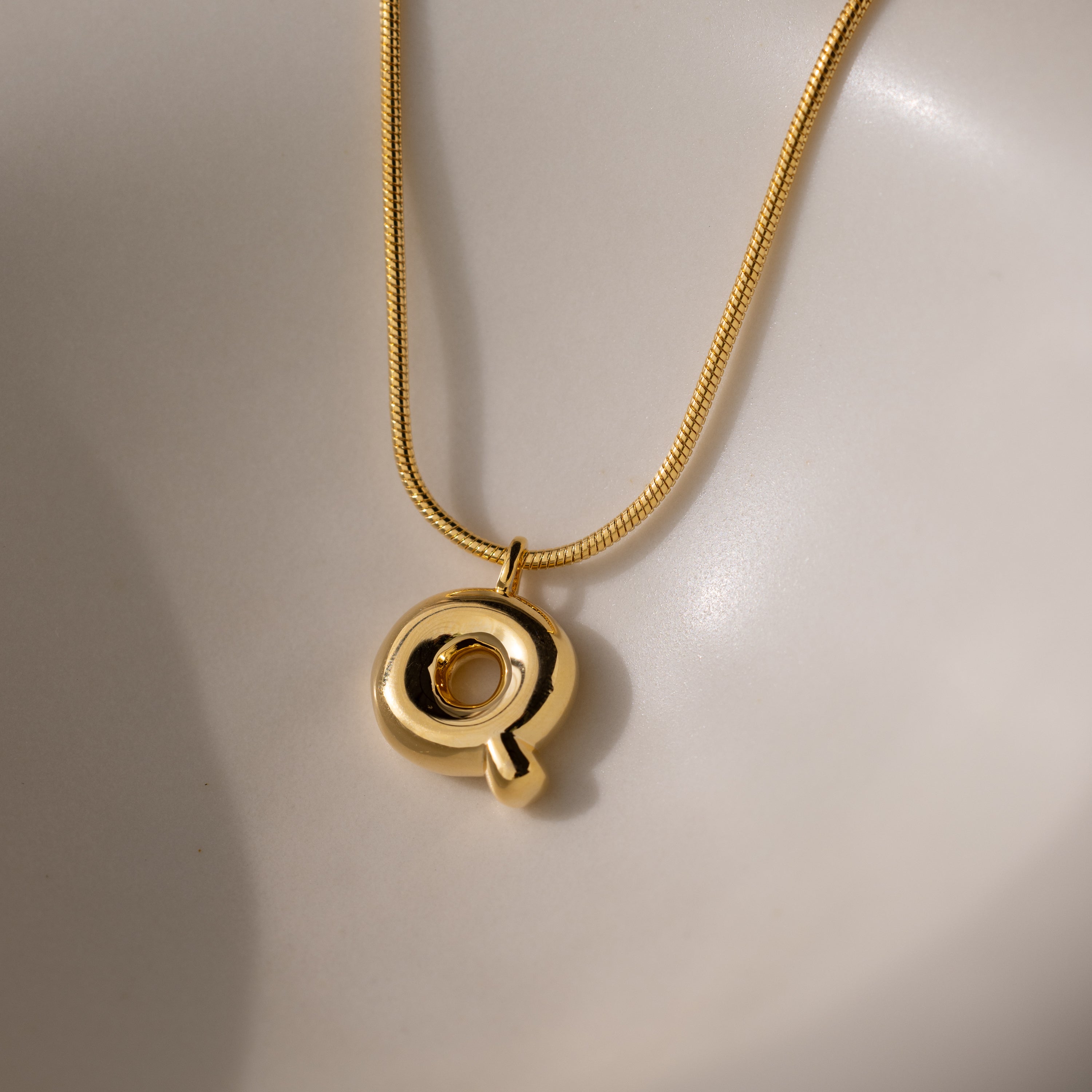 The Bubble Letter Necklace in 18K Gold features a circular, personalized pendant and is displayed on a smooth, light-colored surface.