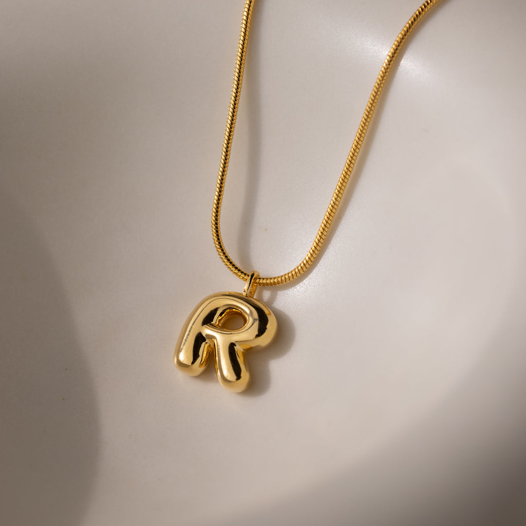 The Bubble Letter Necklace in 18K Gold features a shiny, chunky letter R pendant with a bold bubble design, displayed on a smooth, light surface.