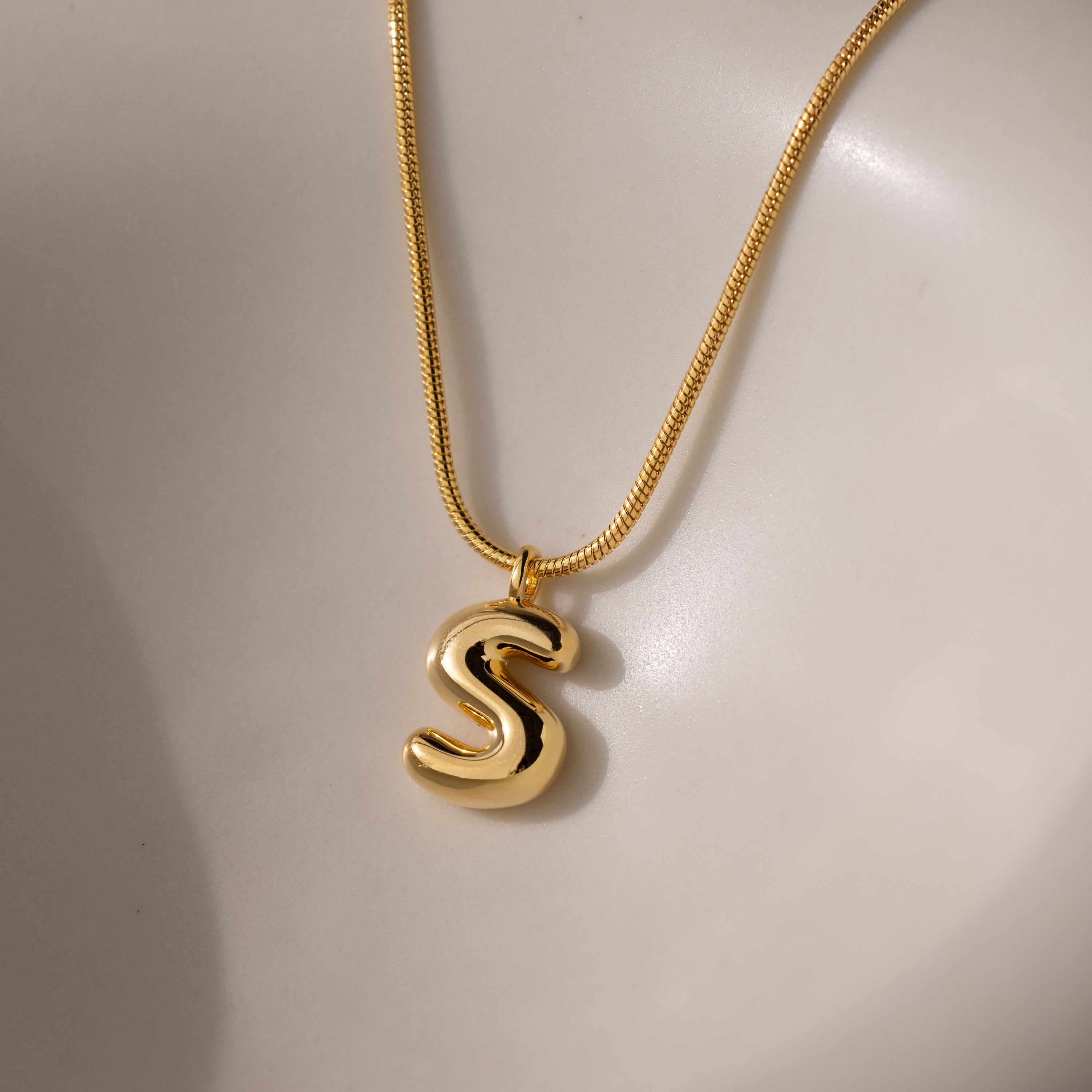The Bubble Letter Necklace in 18K Gold features a shiny, chunky letter S pendant on a smooth, light-colored surface—perfect for lovers of personalized jewelry.
