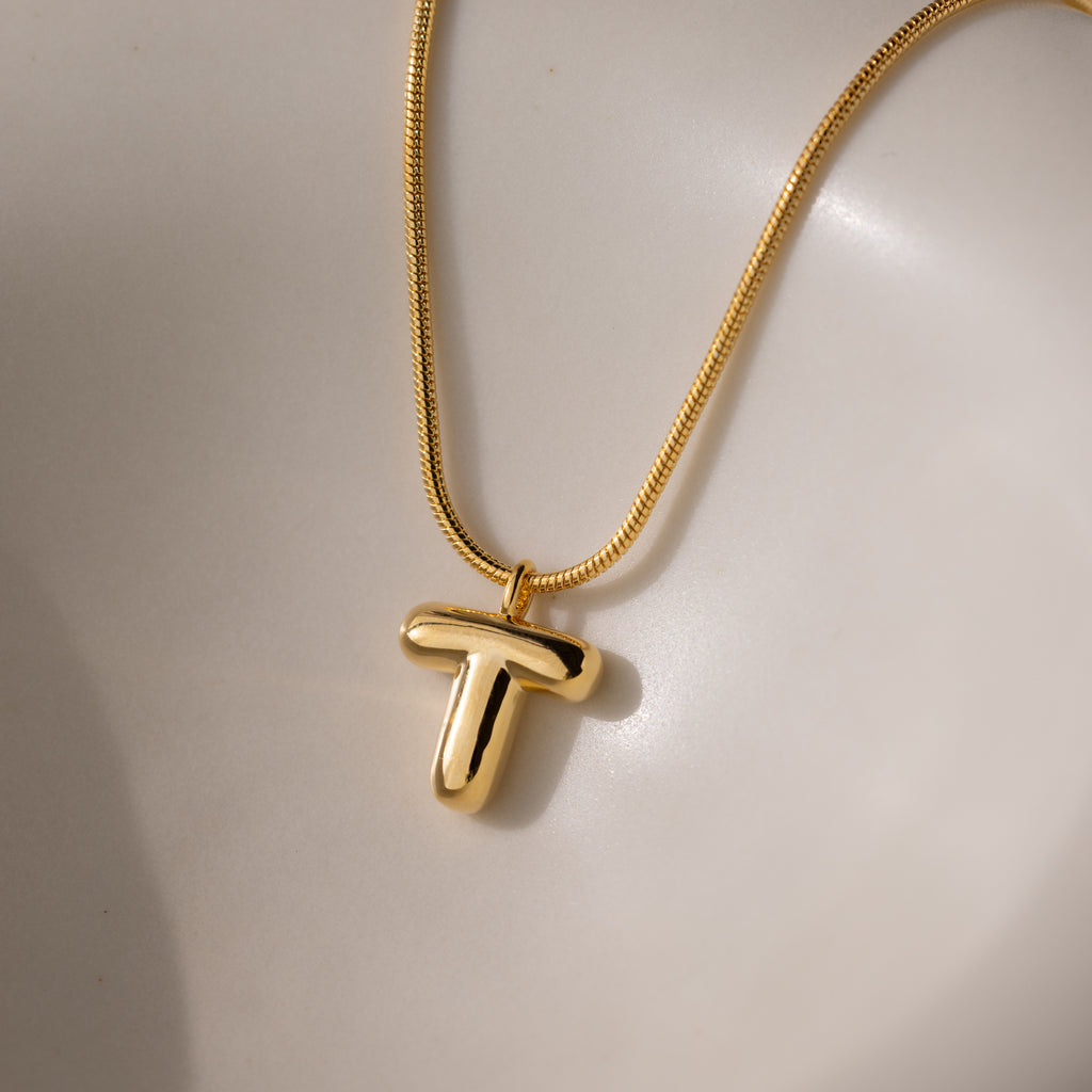 The Bubble Letter Necklace in 18K Gold features a shiny, bold letter T pendant in a chunky charm style, displayed on a smooth, light surface.
