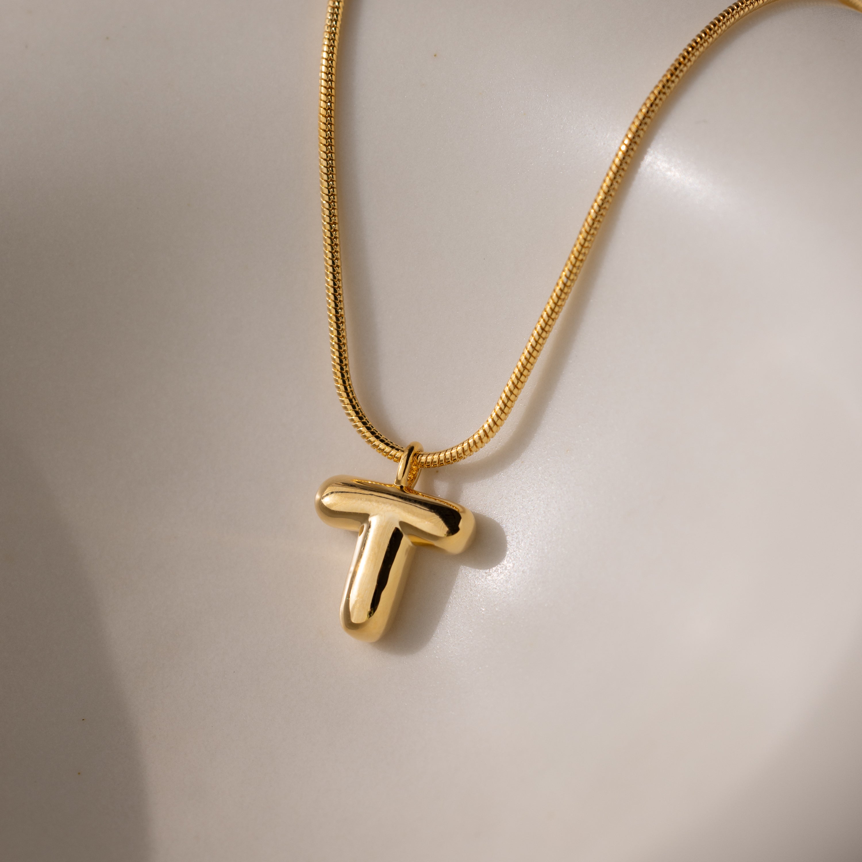 The Bubble Letter Necklace in 18K Gold features a shiny, bold letter T pendant in a chunky charm style, displayed on a smooth, light surface.