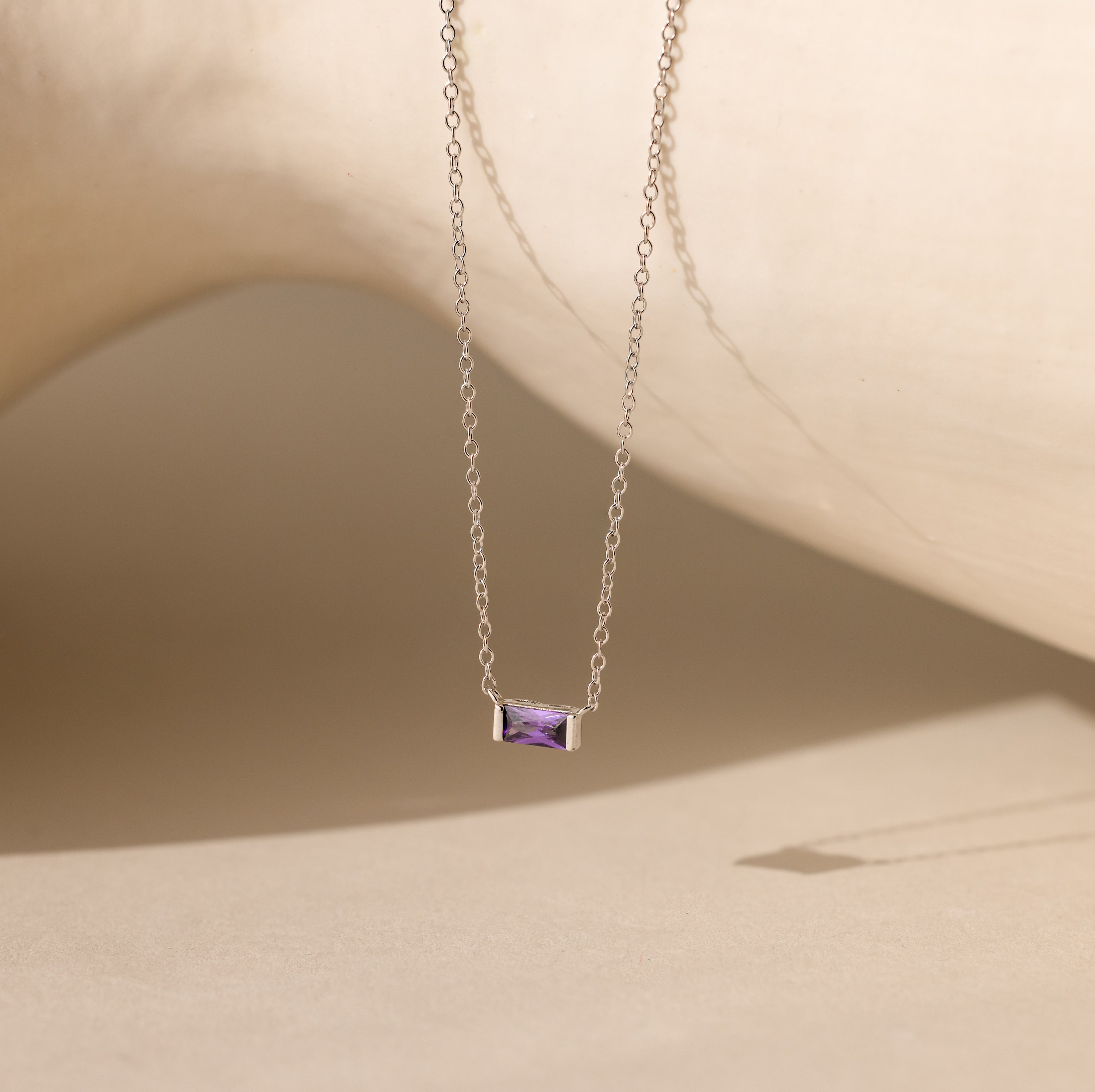 A silver Emery Birthstone Necklace featuring a rectangular amethyst gemstone, styled against a neutral-toned backdrop.