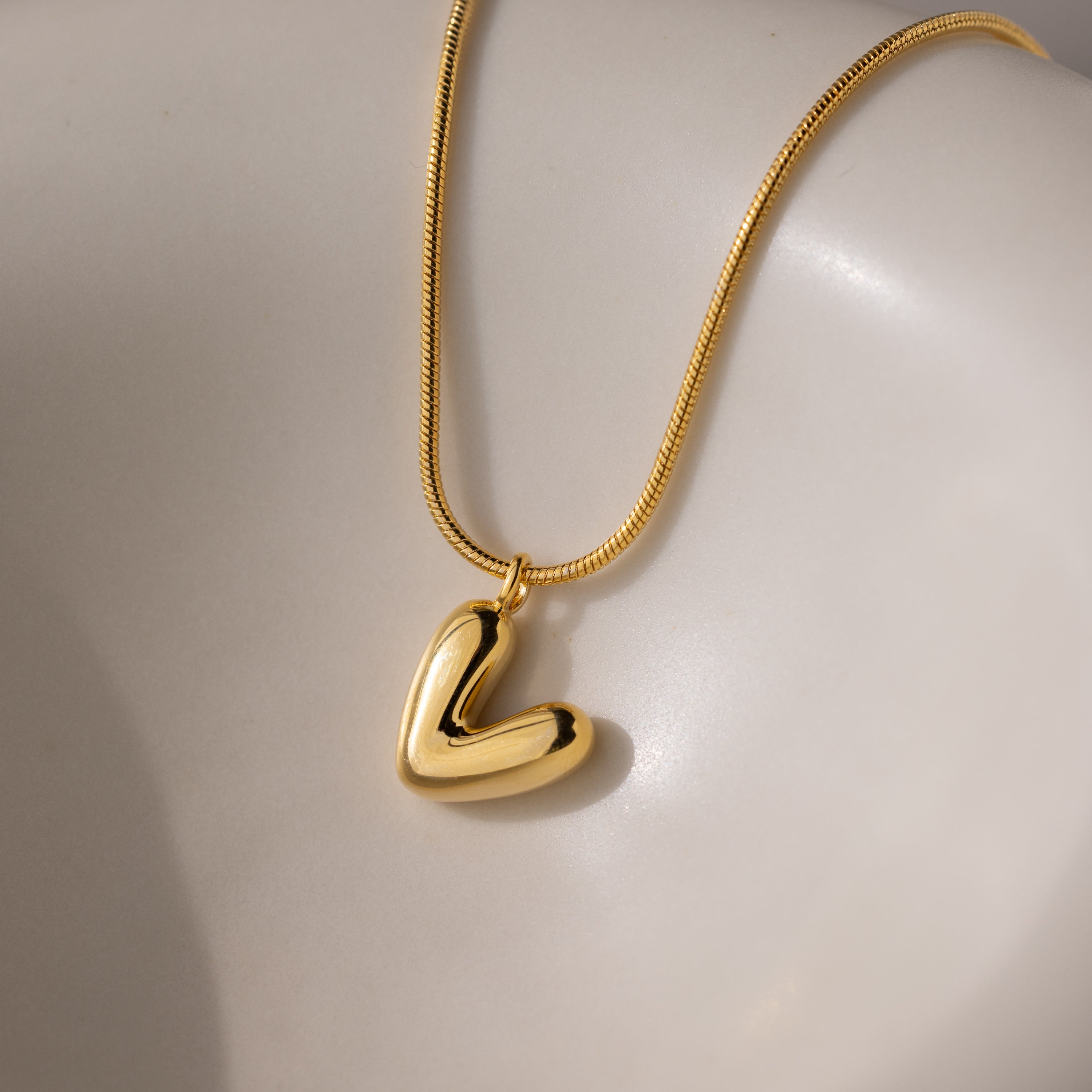 The Bubble Letter Necklace in 18K Gold, featuring a shiny abstract heart-shaped pendant and chunky letter charm, rests on a smooth, light-colored surface.