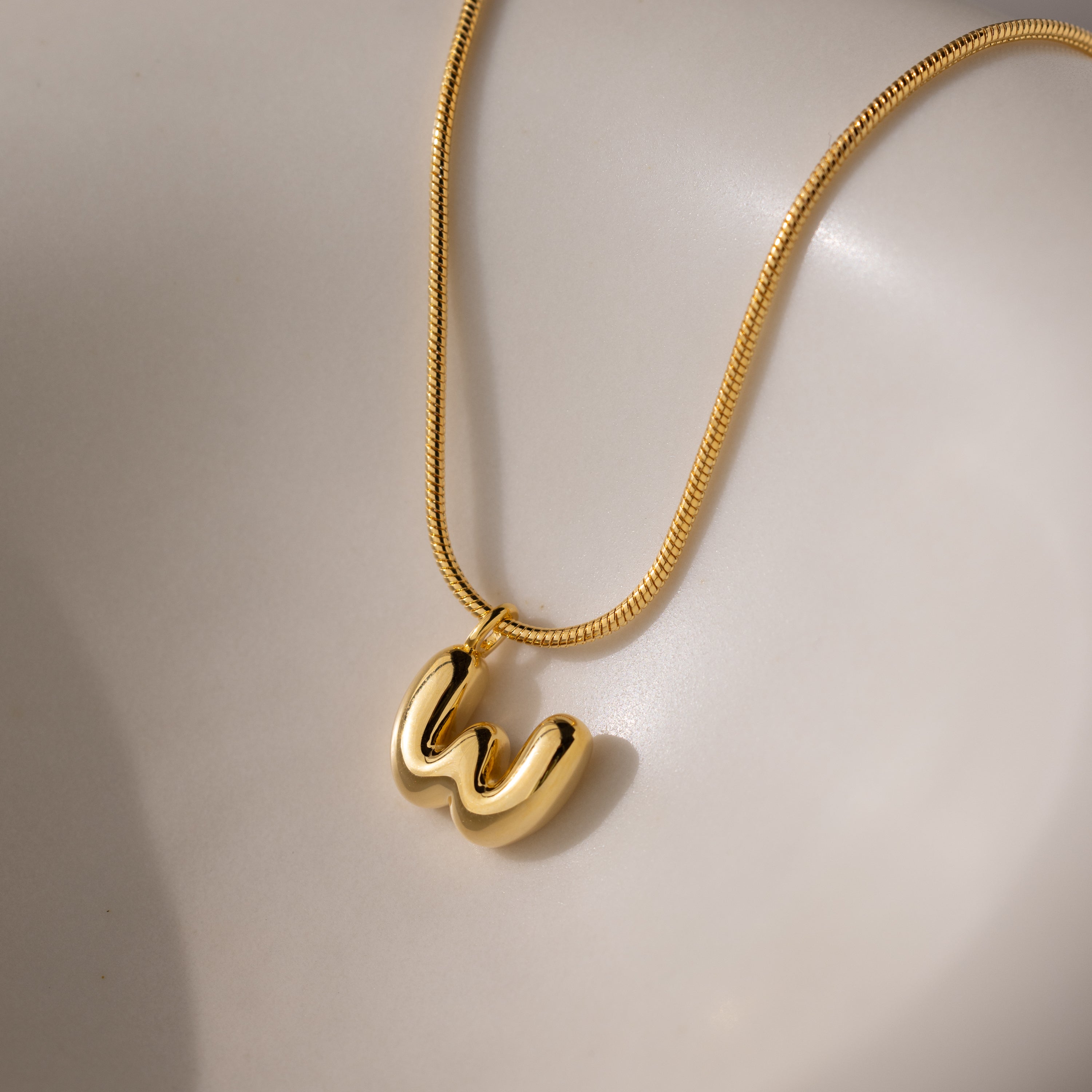 The Bubble Letter Necklace in 18K Gold features a shiny "W" pendant, elegantly displayed on a smooth, light-colored surface.