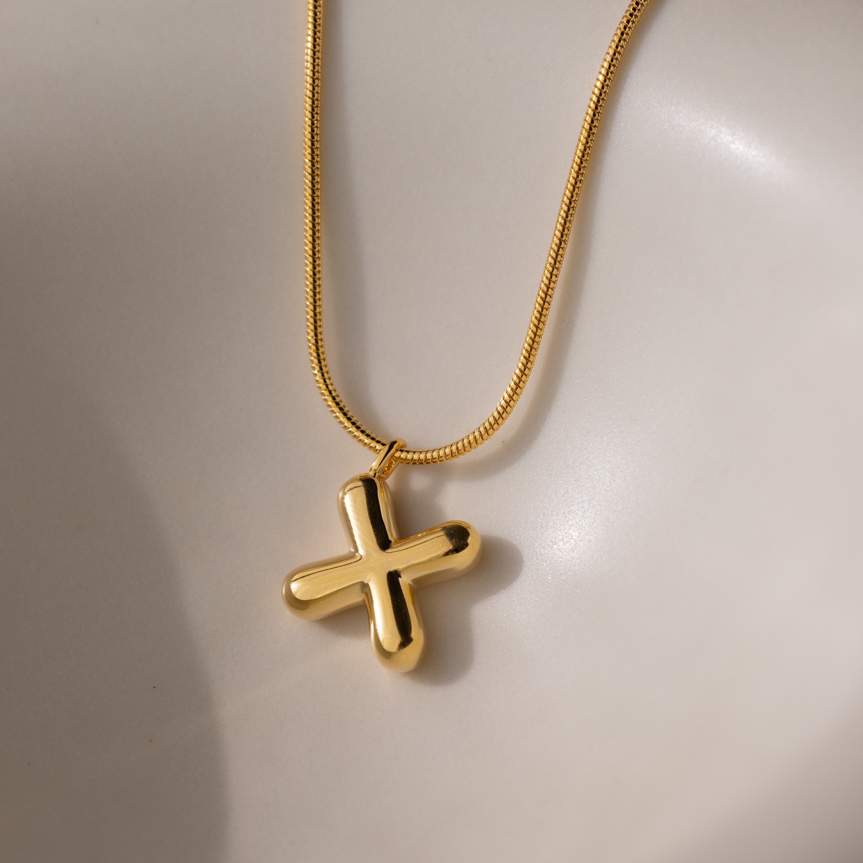 The Bubble Letter Necklace in 18K Gold features a chunky letter charm and a gold cross, displayed on a white surface.