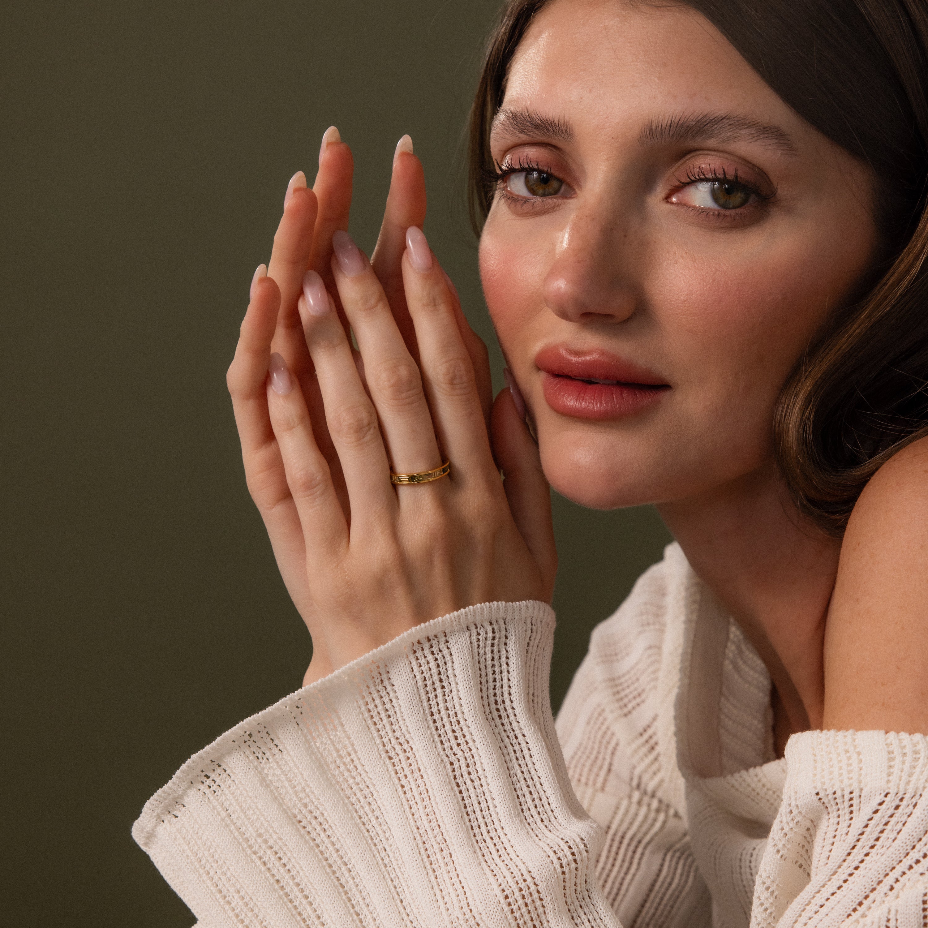 Woman with long brown hair wears a white sweater and the Peridot Inlay Eternity Ring, posing with hands together against a green background.
