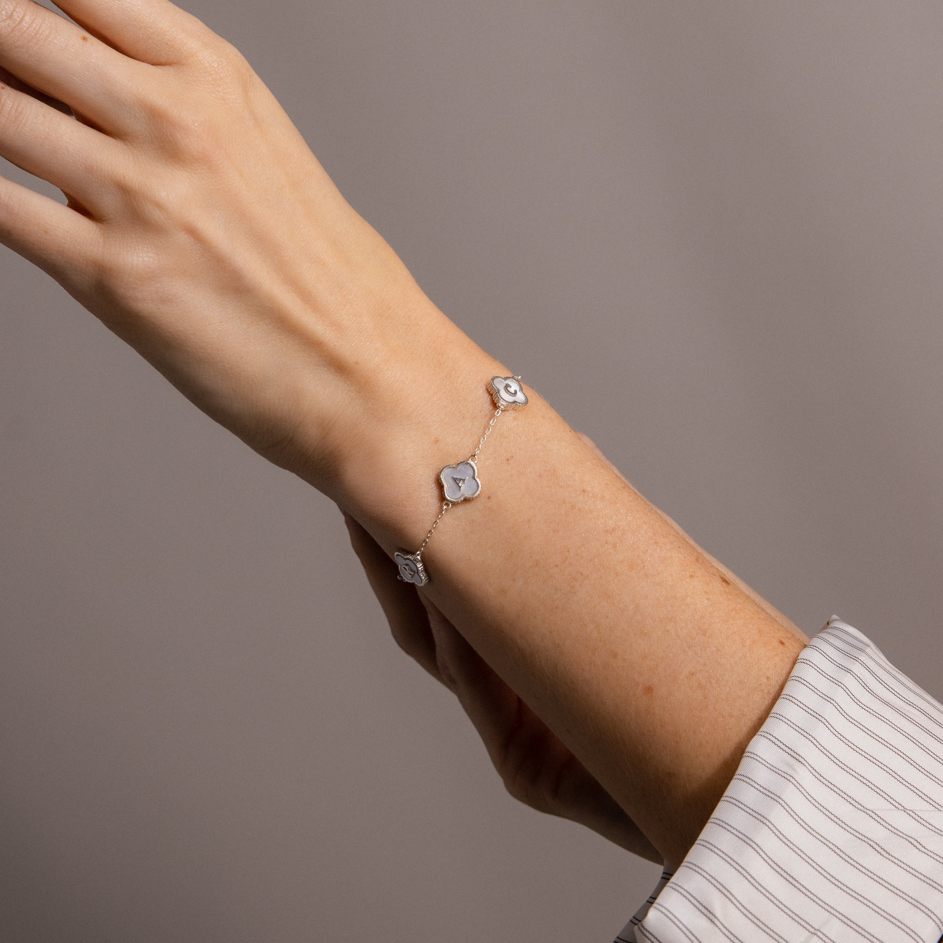 A hand shows off the Pearl Clover Letter Bracelet, featuring three mother of pearl clovers on a delicate silver chain, against a neutral background.
