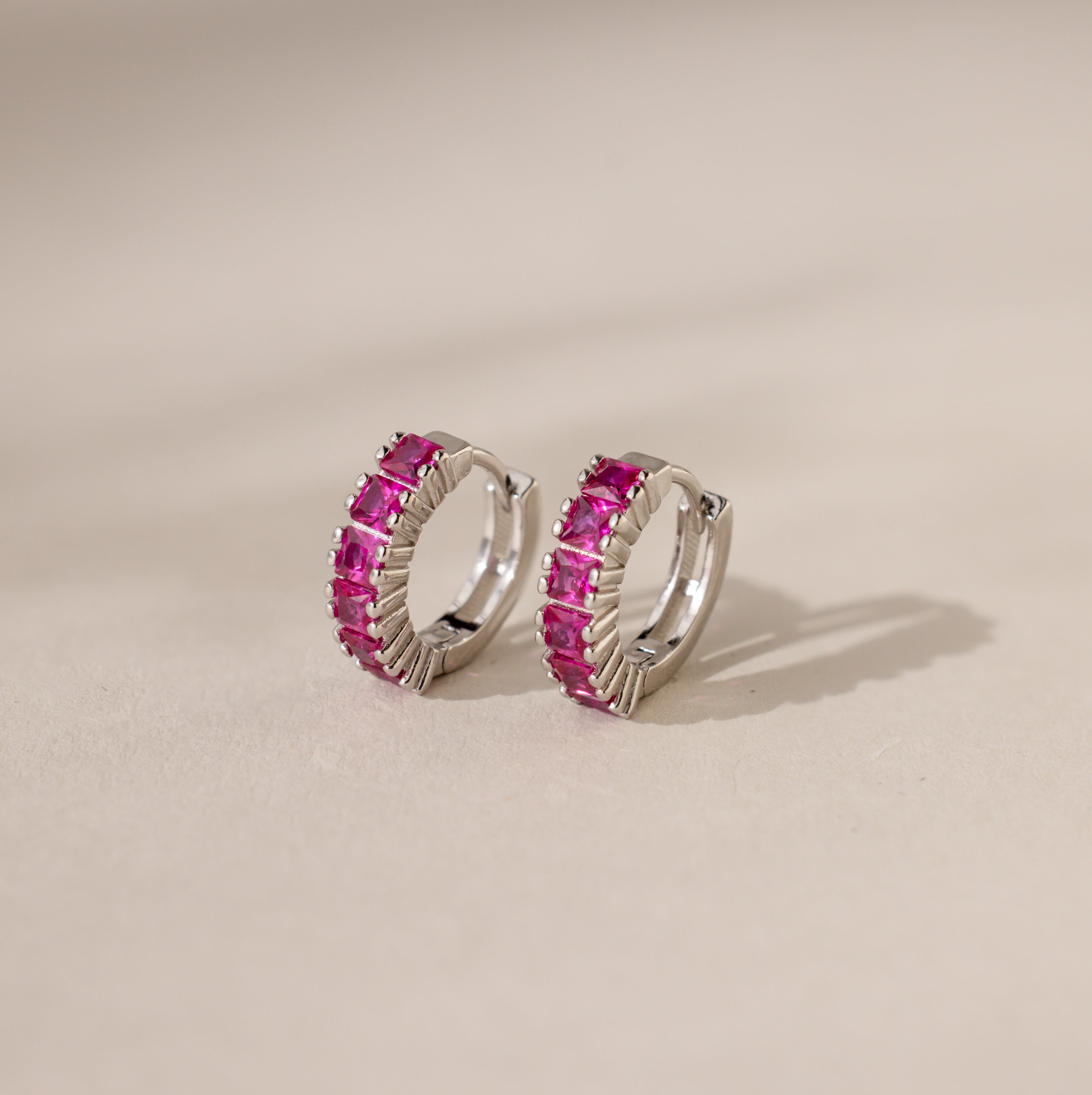 Genevieve Ruby Huggies: silver huggie hoops featuring a row of pink gemstones, displayed on a light beige surface—an elegant choice for birthstone earrings.