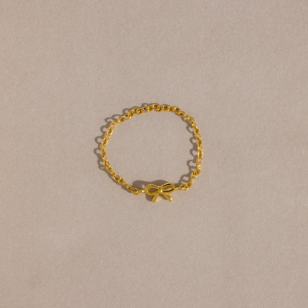 A delicate gold chain ring with a small, polished bow charm placed neatly on a soft beige background.
