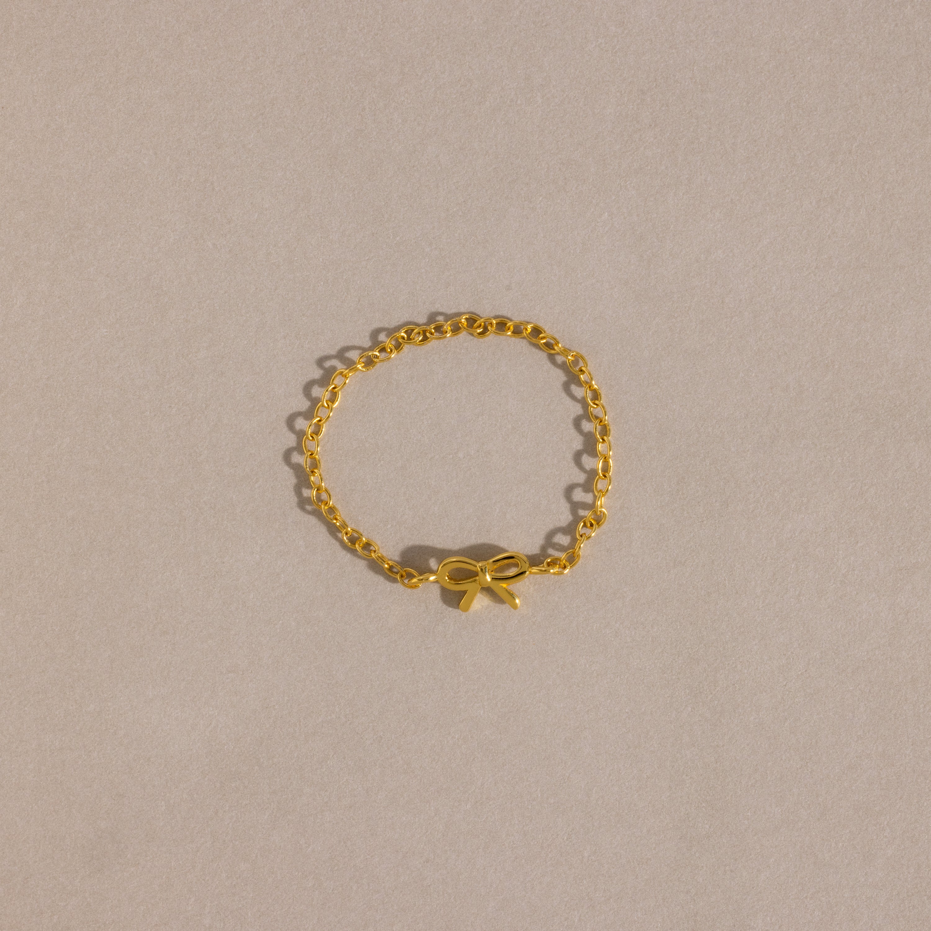 A delicate gold chain ring with a small, polished bow charm placed neatly on a soft beige background.