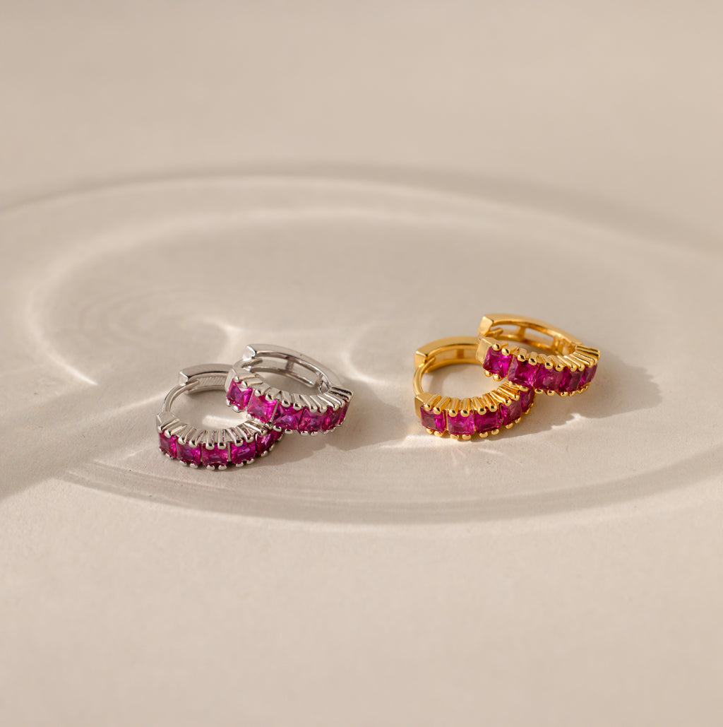 Pairs of Genevieve Birthstone Huggies shown in silver and gold finishes, each set with vivid ruby gemstones, displayed on a neutral surface with soft light reflections.