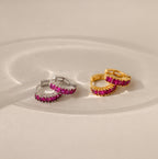 Pairs of Genevieve Birthstone Huggies shown in silver and gold finishes, each set with vivid ruby gemstones, displayed on a neutral surface with soft light reflections.