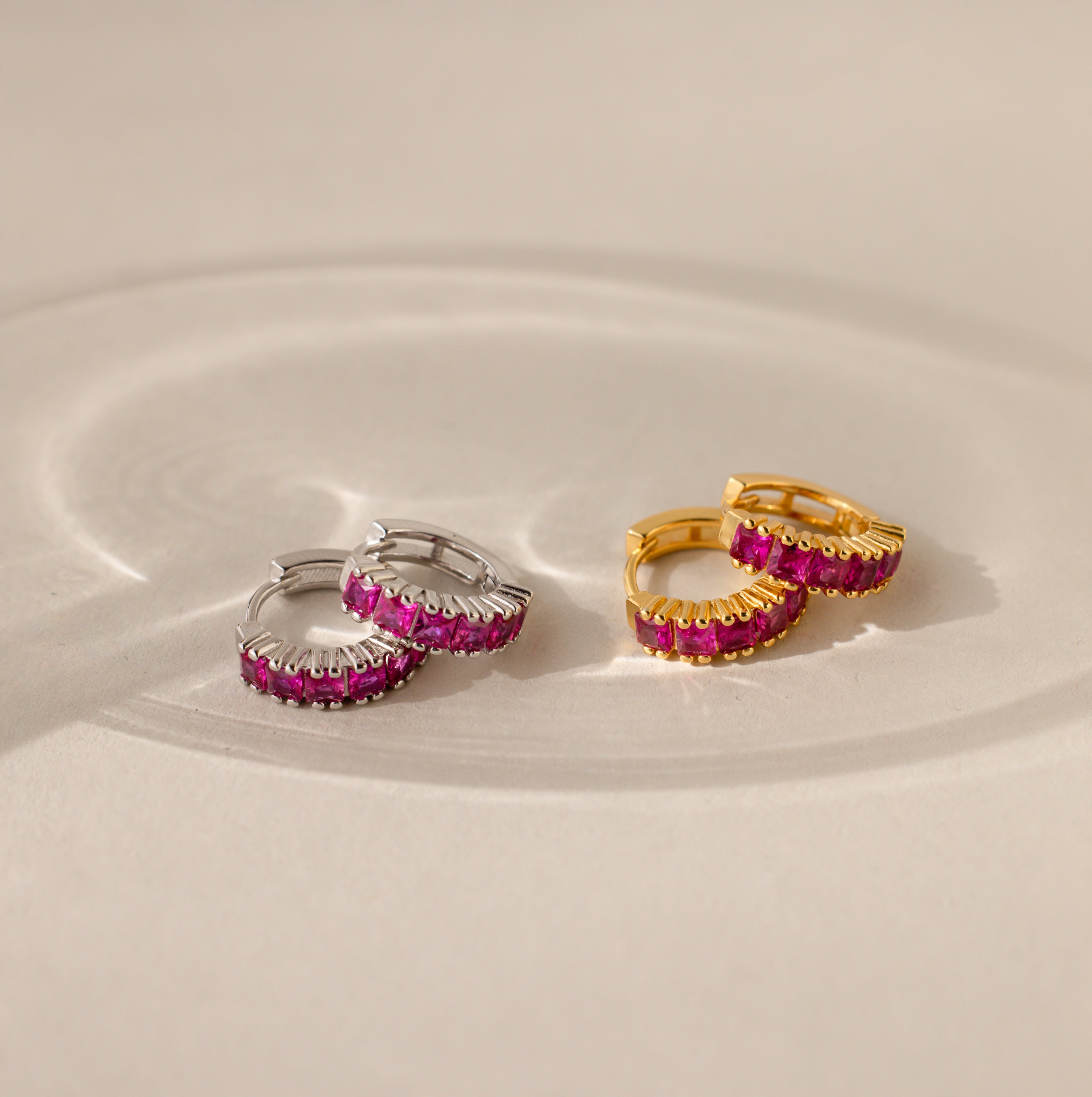 Pairs of Genevieve Birthstone Huggies shown in silver and gold finishes, each set with vivid ruby gemstones, displayed on a neutral surface with soft light reflections.