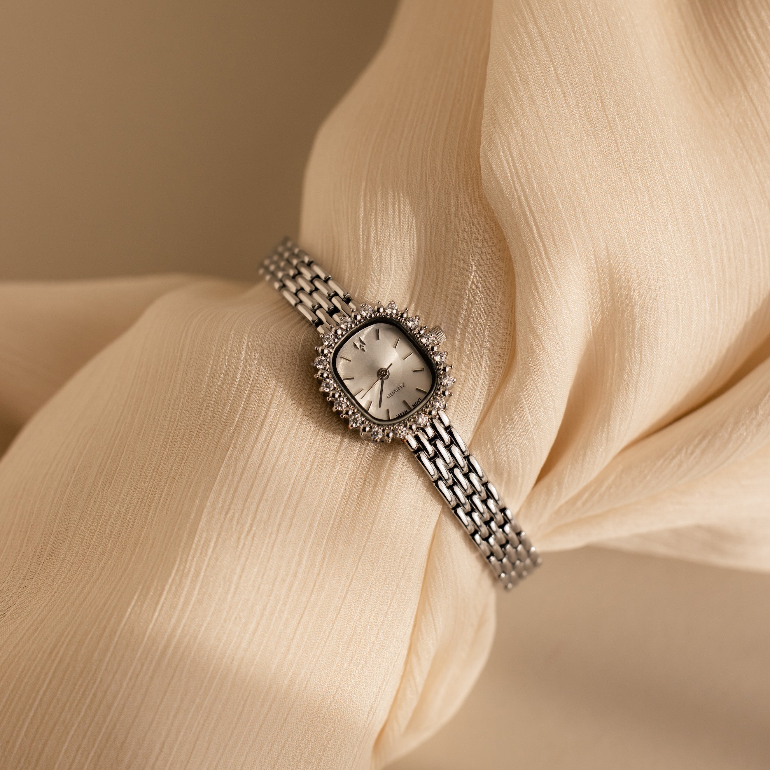 The Pave Blossom Timepiece is an elegant womens watch with a round face and metal band, showcased on soft cream-colored fabric for timeless appeal.