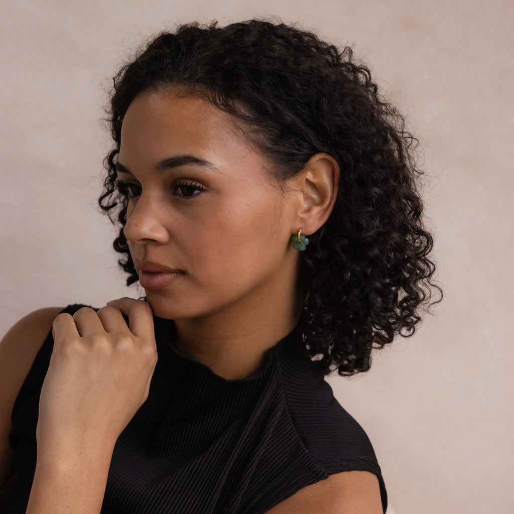 Woman with curly hair wearing Jade Flower Hoops and a black top, looking to the side with her hand near her chin.