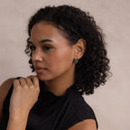 Woman with curly hair wearing Jade Flower Hoops and a black top, looking to the side with her hand near her chin.