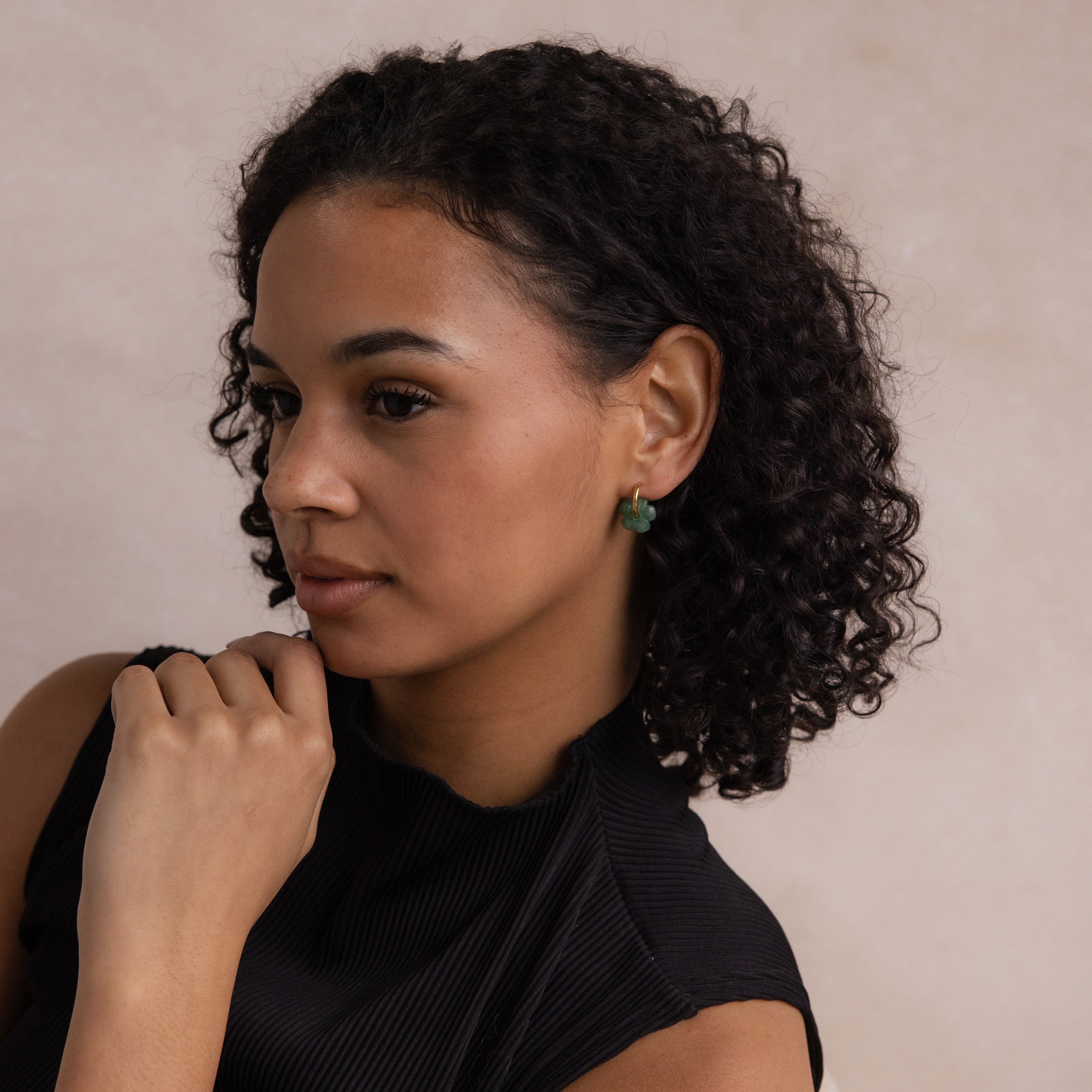 Woman with curly hair wearing Jade Flower Hoops and a black top, looking to the side with her hand near her chin.