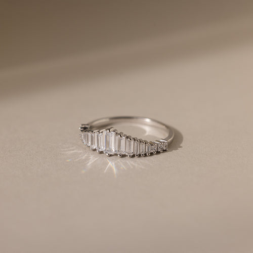 The Art Deco Diamond Ring in Sterling Silver features rectangular diamonds set in a curved line, casting a soft shadow on a beige surface—a striking statement piece.