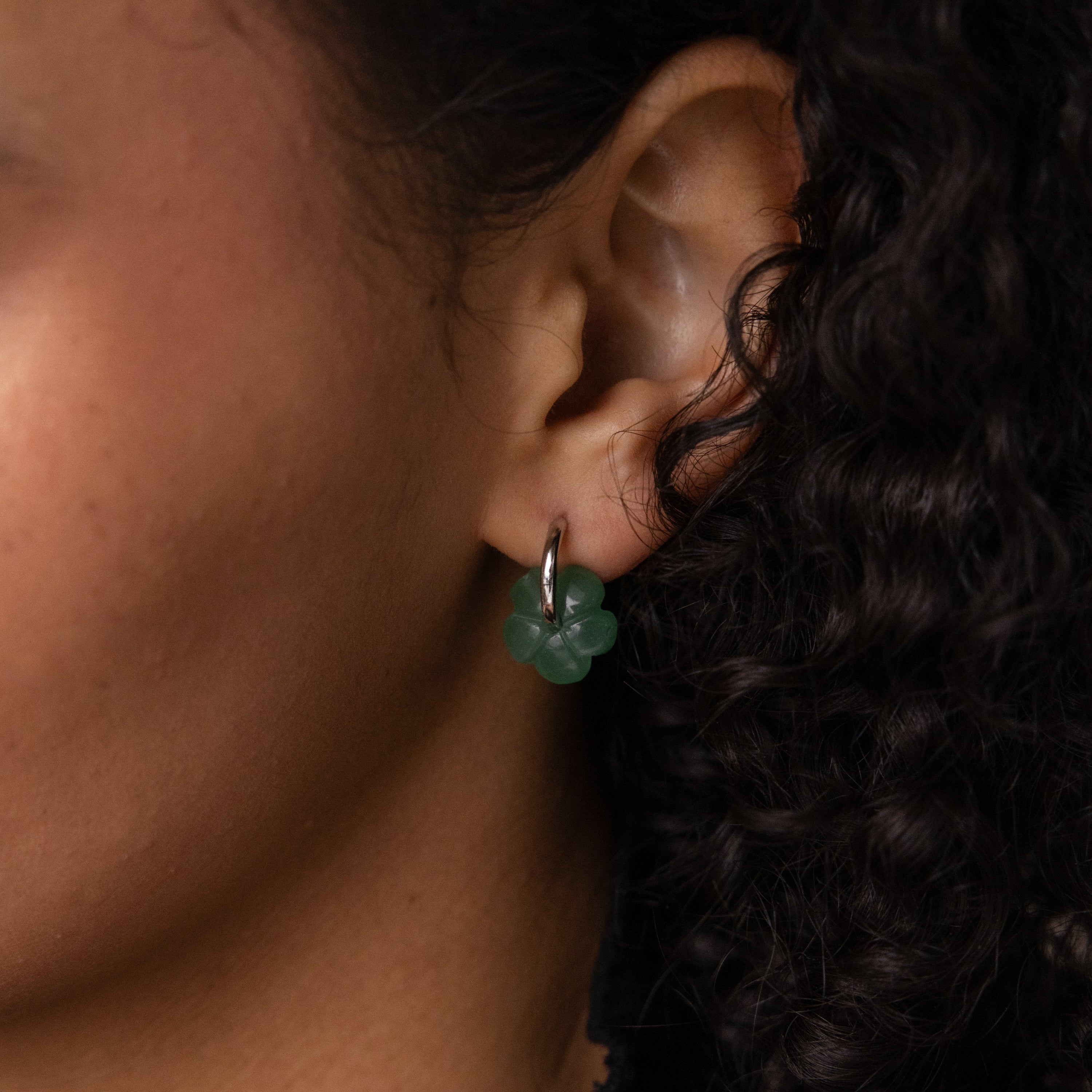 Close-up of an ear wearing Jade Flower Hoops in Silver, with delicate jade flower details and curly dark hair partially visible.