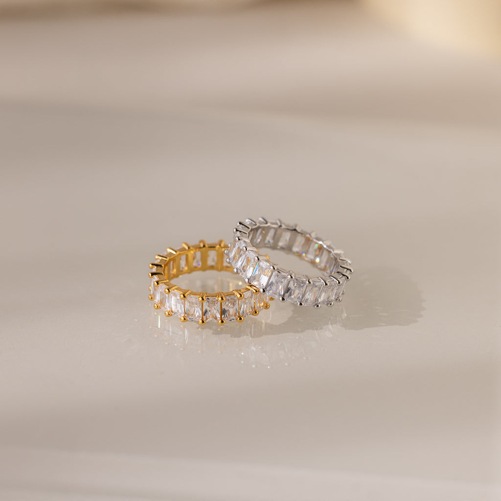The gold statement ring with baguette diamonds and the Colette Eternity Ring, a classic silver design, are displayed on a light, reflective surface.