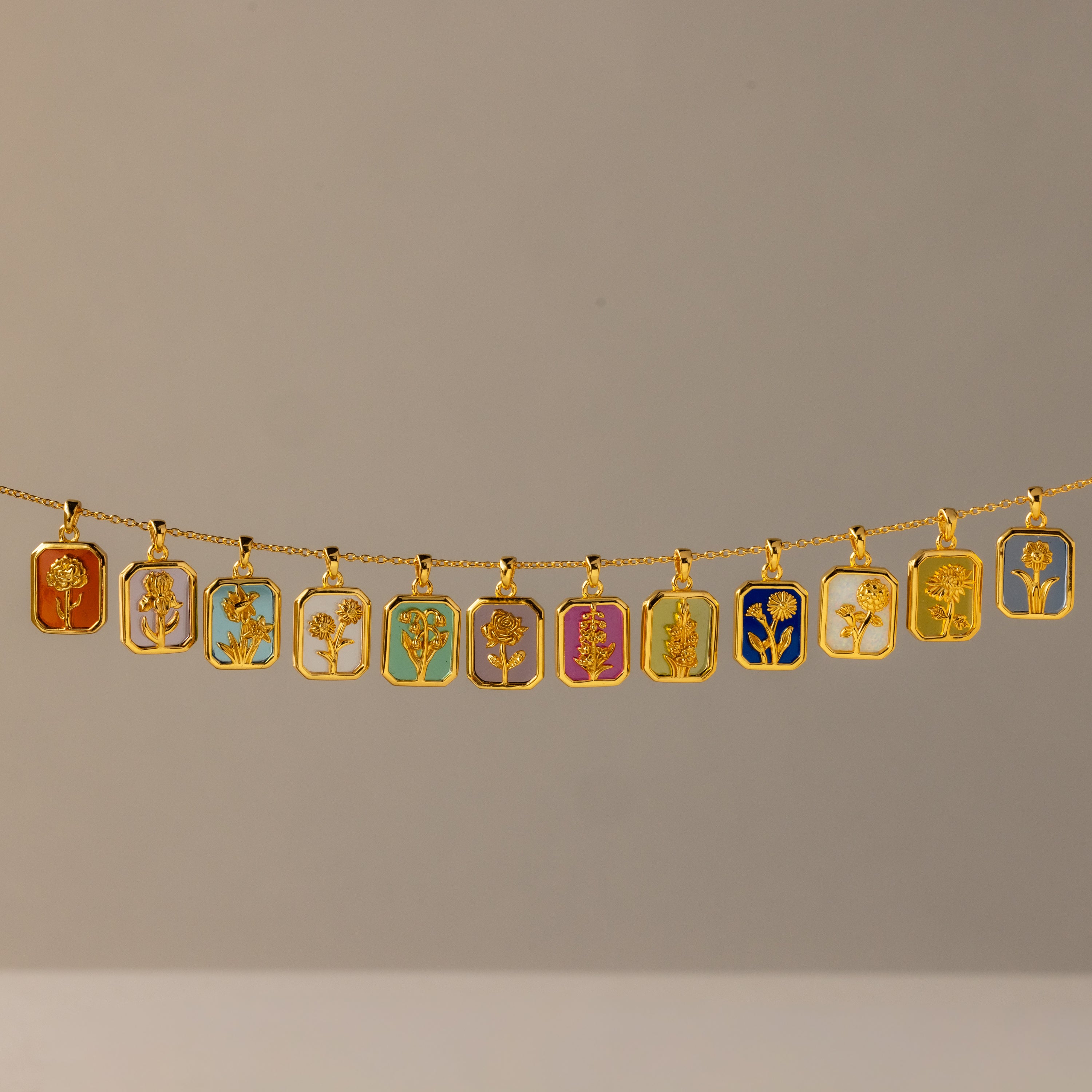 The Birth Flower Charm Necklace features a gold chain with twelve rectangular pendants, each showcasing a distinct colorful floral design—an elegant piece that brings artistic flair to any outfit.