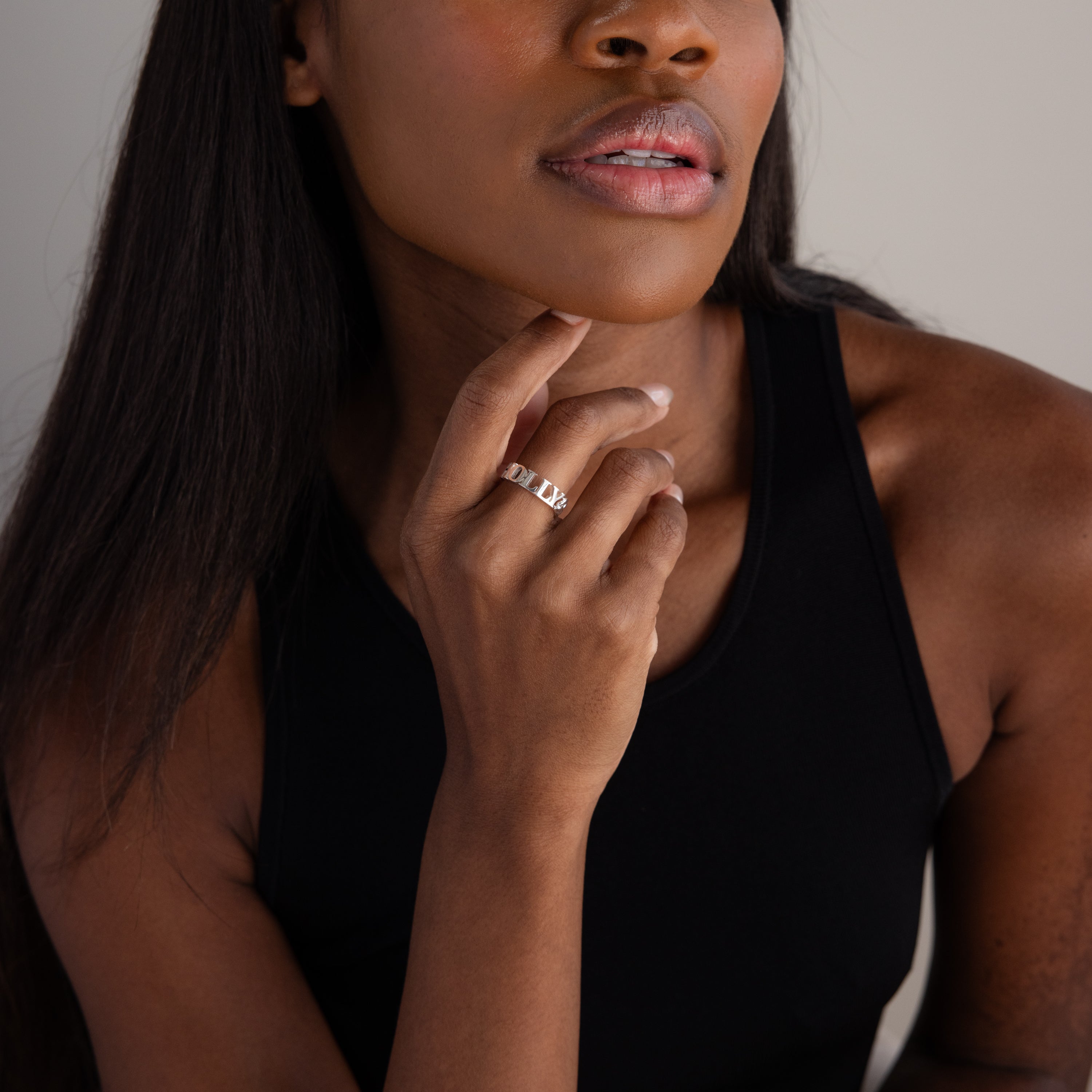 A woman in a black tank top touches her chin, showcasing the Newsletter Curb Name Ring in Sterling Silver with its sleek chain-inspired design on her finger.