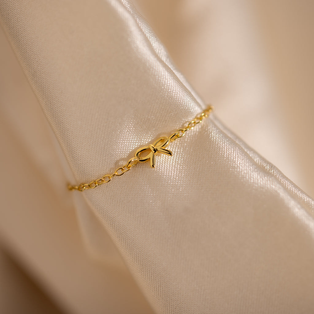 Close-up image of a delicate gold chain ring with a small bow charm in the center, elegantly draped over a folded satin fabric.