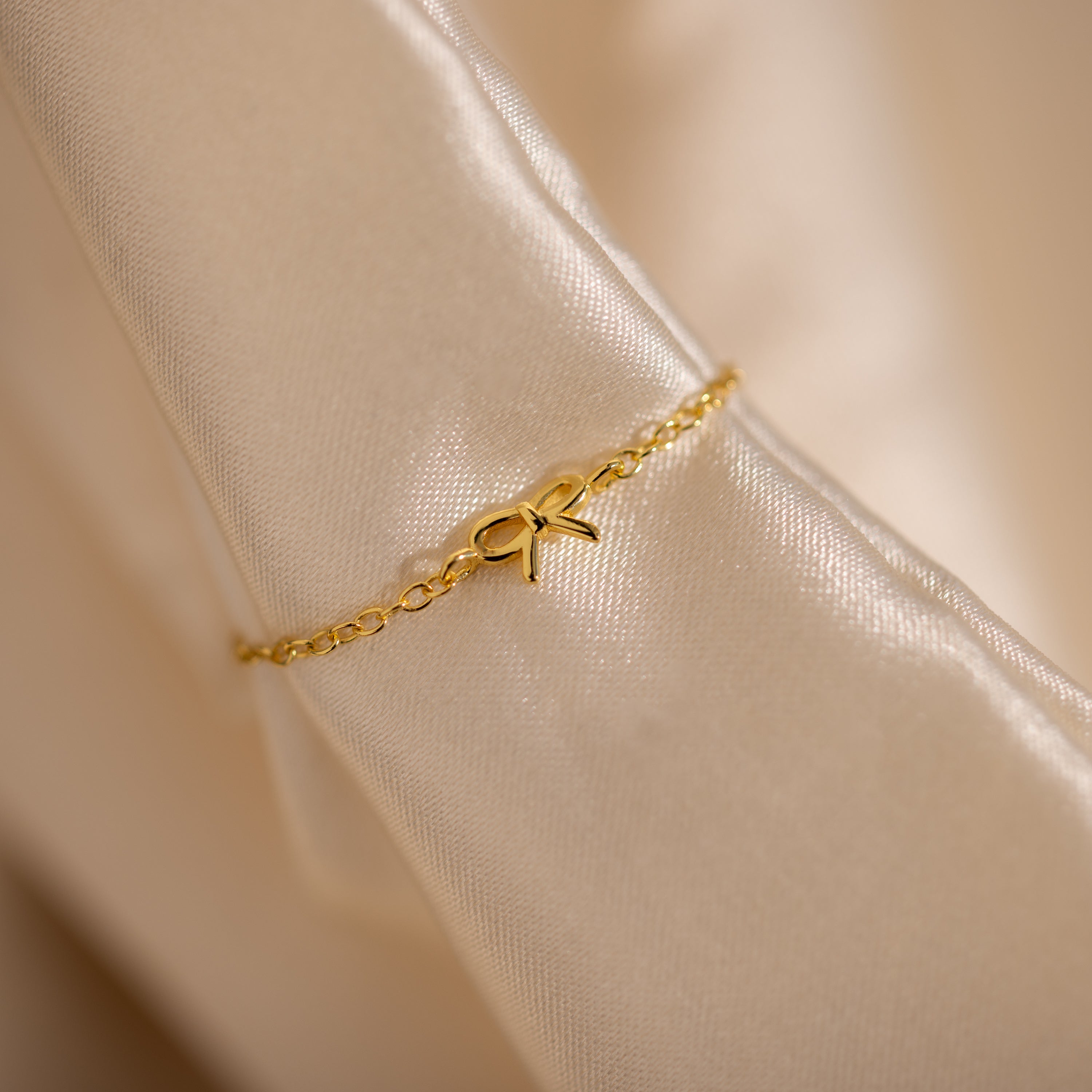 Close-up image of a delicate gold chain ring with a small bow charm in the center, elegantly draped over a folded satin fabric.