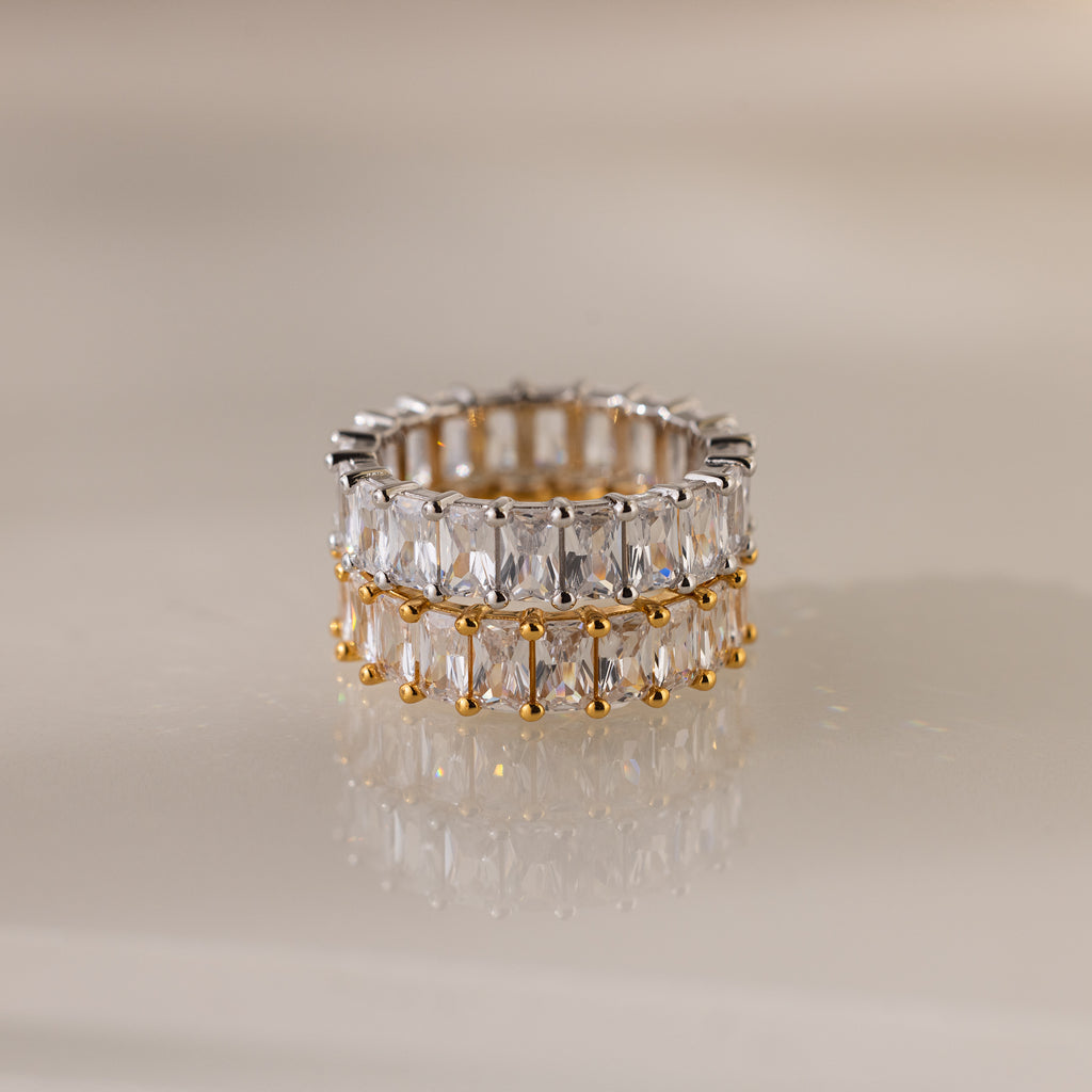 The Colette Eternity Ring, crafted in gold and silver with two rows of rectangular baguette diamonds, is displayed on a reflective beige surface.