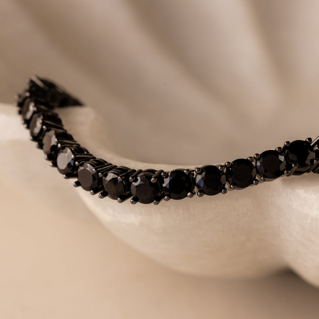 A close-up of the Onyx Tennis Bracelet, featuring black CZ diamonds, displayed on a white textured surface.