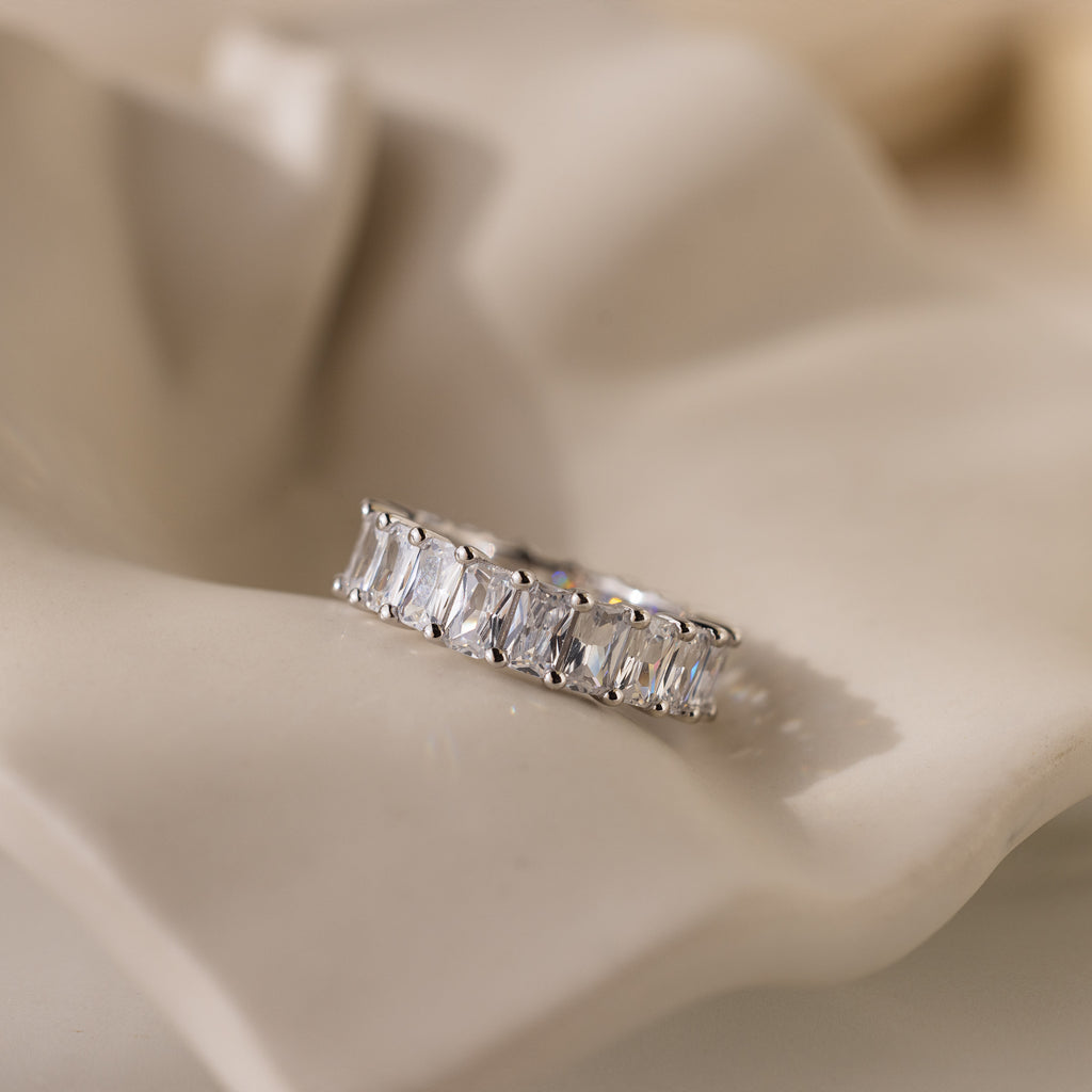 The Colette Eternity Ring, featuring rectangular baguette diamonds in silver, rests on a soft cream-colored fabric.