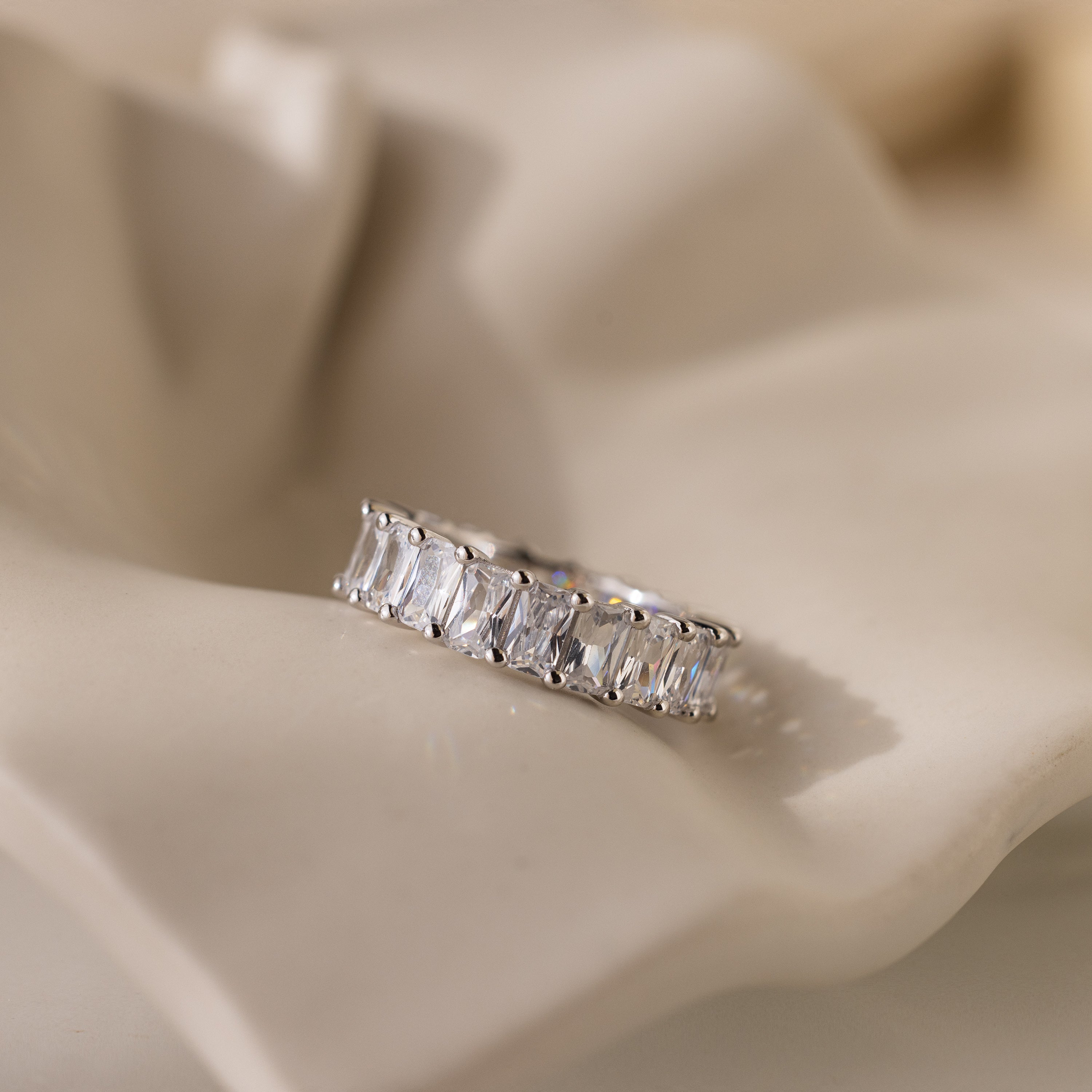 The Colette Eternity Ring, featuring rectangular baguette diamonds in silver, rests on a soft cream-colored fabric.