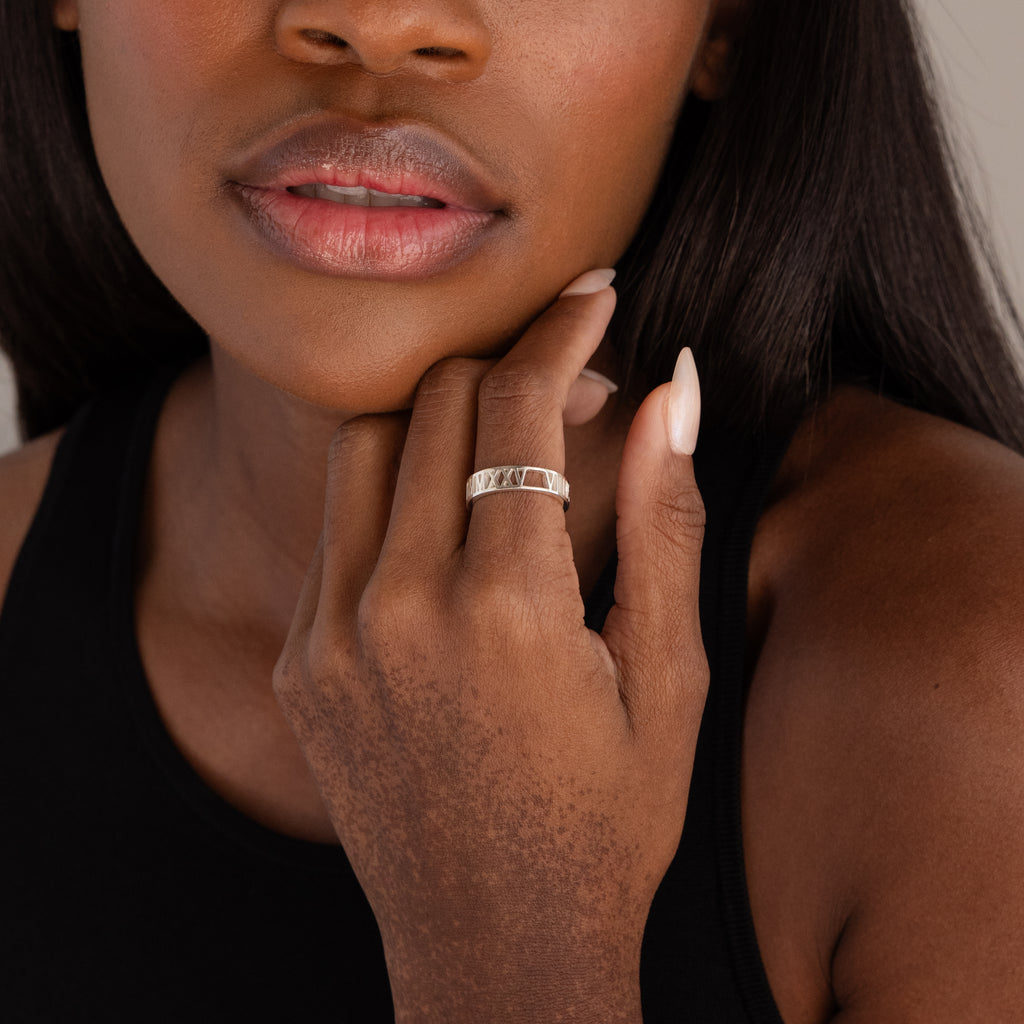 A woman touches her face, revealing her lips and part of her black top while wearing the Roman Numerals Ring in Sterling Silver.