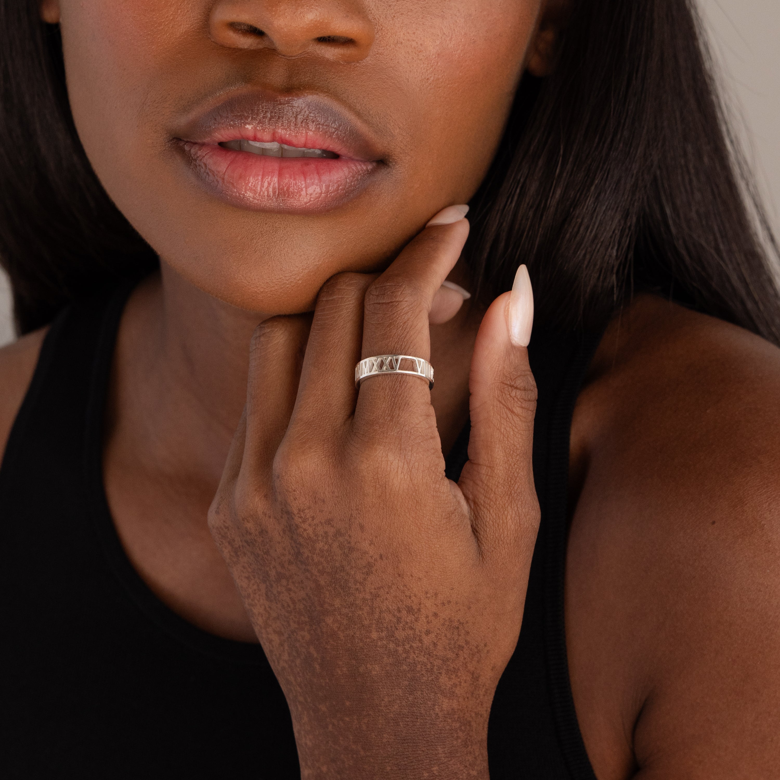 A woman touches her face, revealing her lips and part of her black top while wearing the Roman Numerals Ring in Sterling Silver.