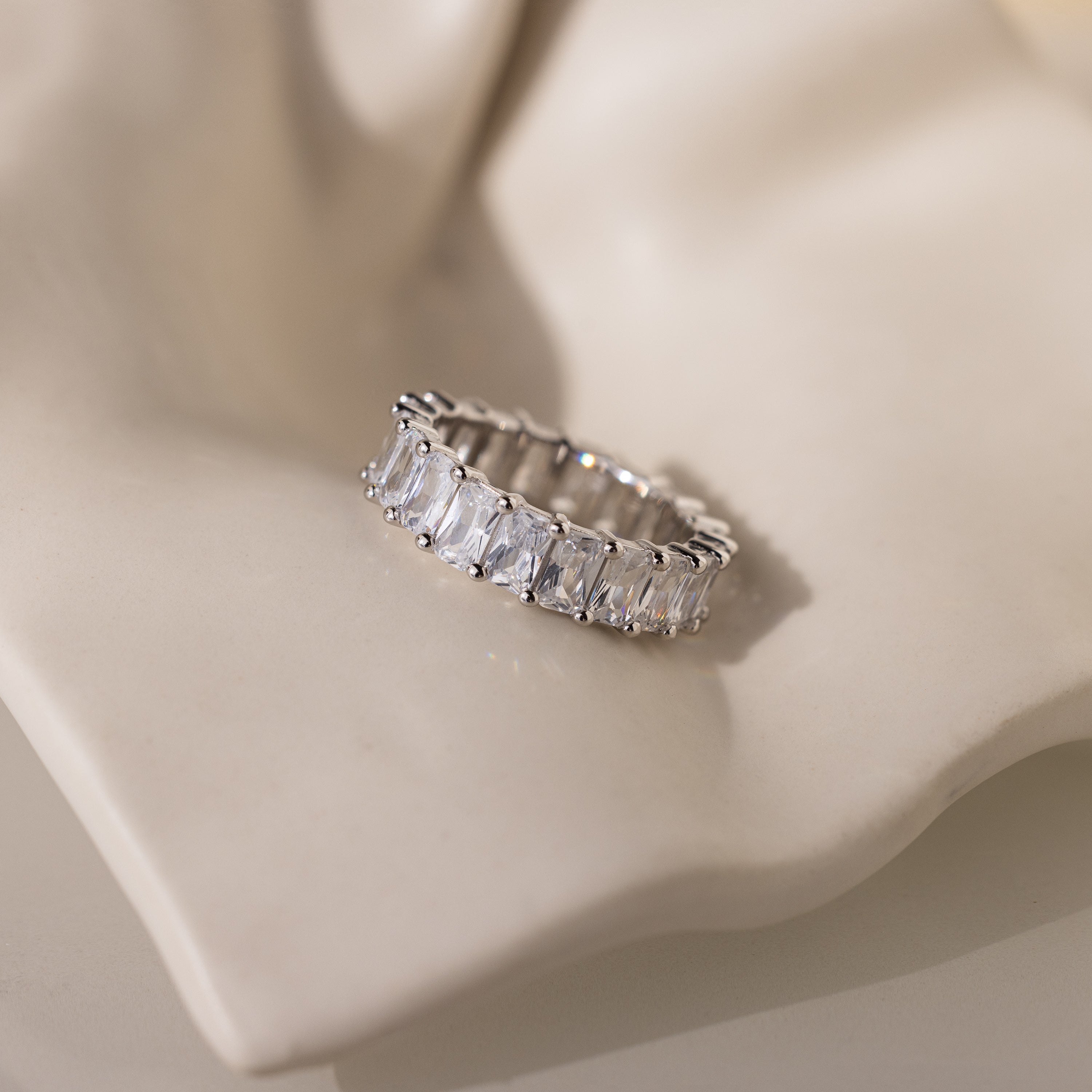 The Colette Eternity Ring, featuring silver and rectangular baguette diamonds, rests on a soft cream-colored textured surface and radiates subtle opulence.