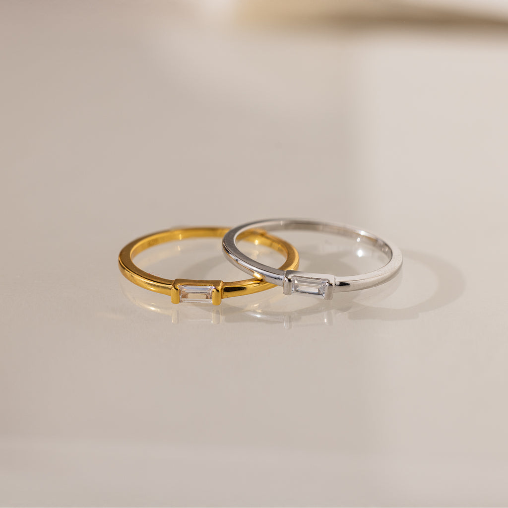 Two Baguette Diamond Rings, one gold and one silver, each with a slim band and small rectangular diamond in an east-west setting, rest on a light surface.