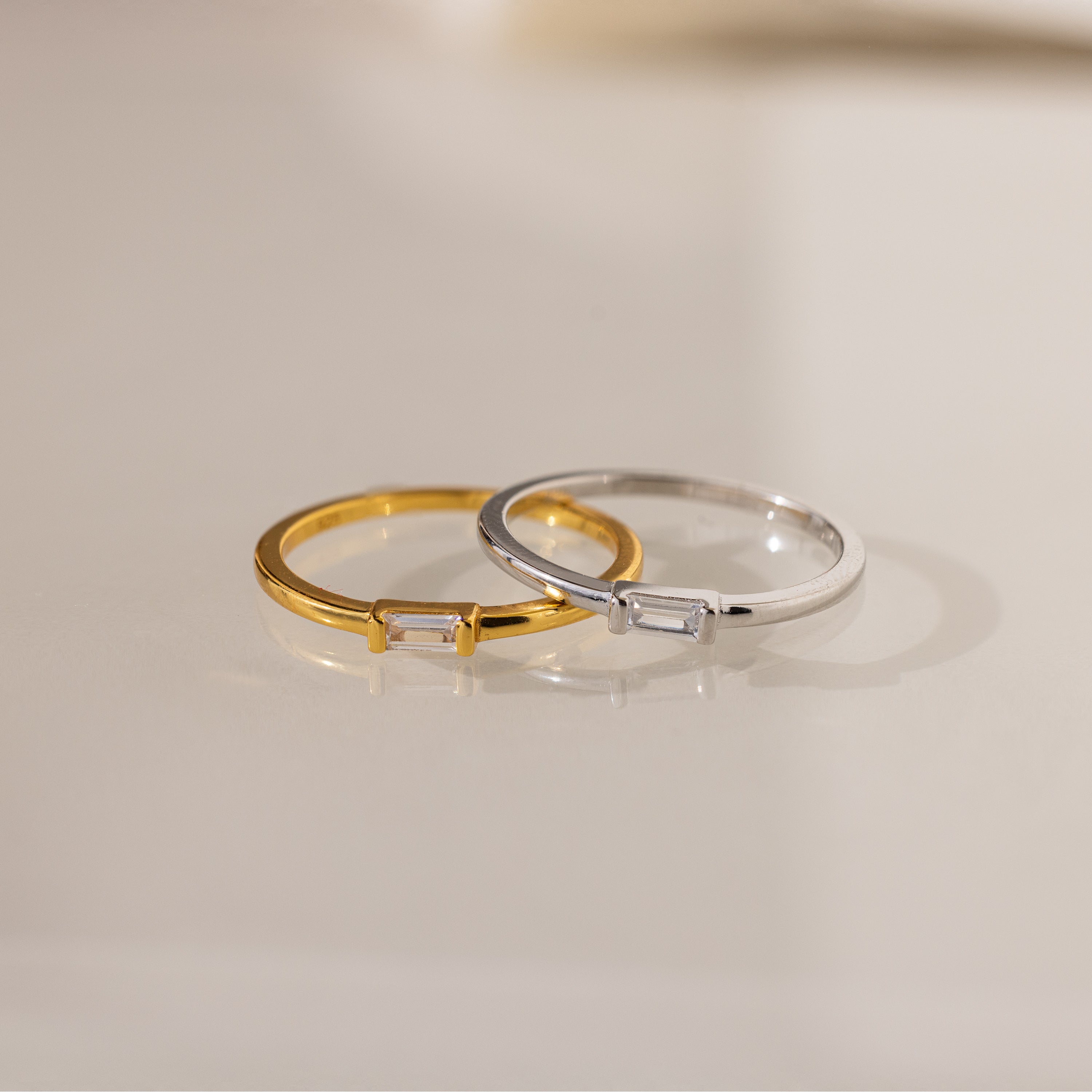 Two Baguette Diamond Rings, one gold and one silver, each with a slim band and small rectangular diamond in an east-west setting, rest on a light surface.